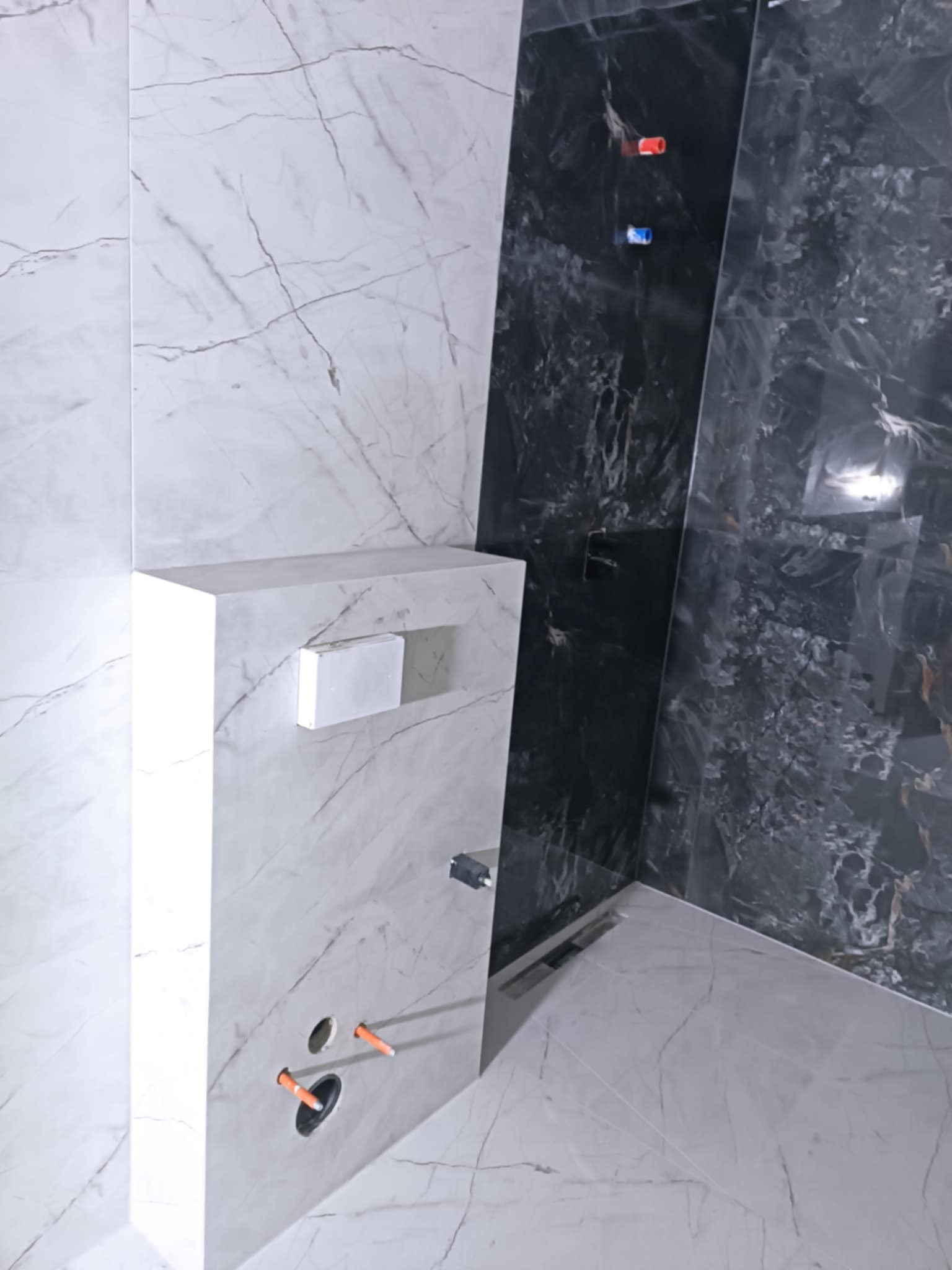 Nowoczesna łazienka w trakcie wykańczania: białe i czarne płytki imitujące marmur, zabudowa WC z półką, widoczne otwory na armaturę i odpływ liniowy.