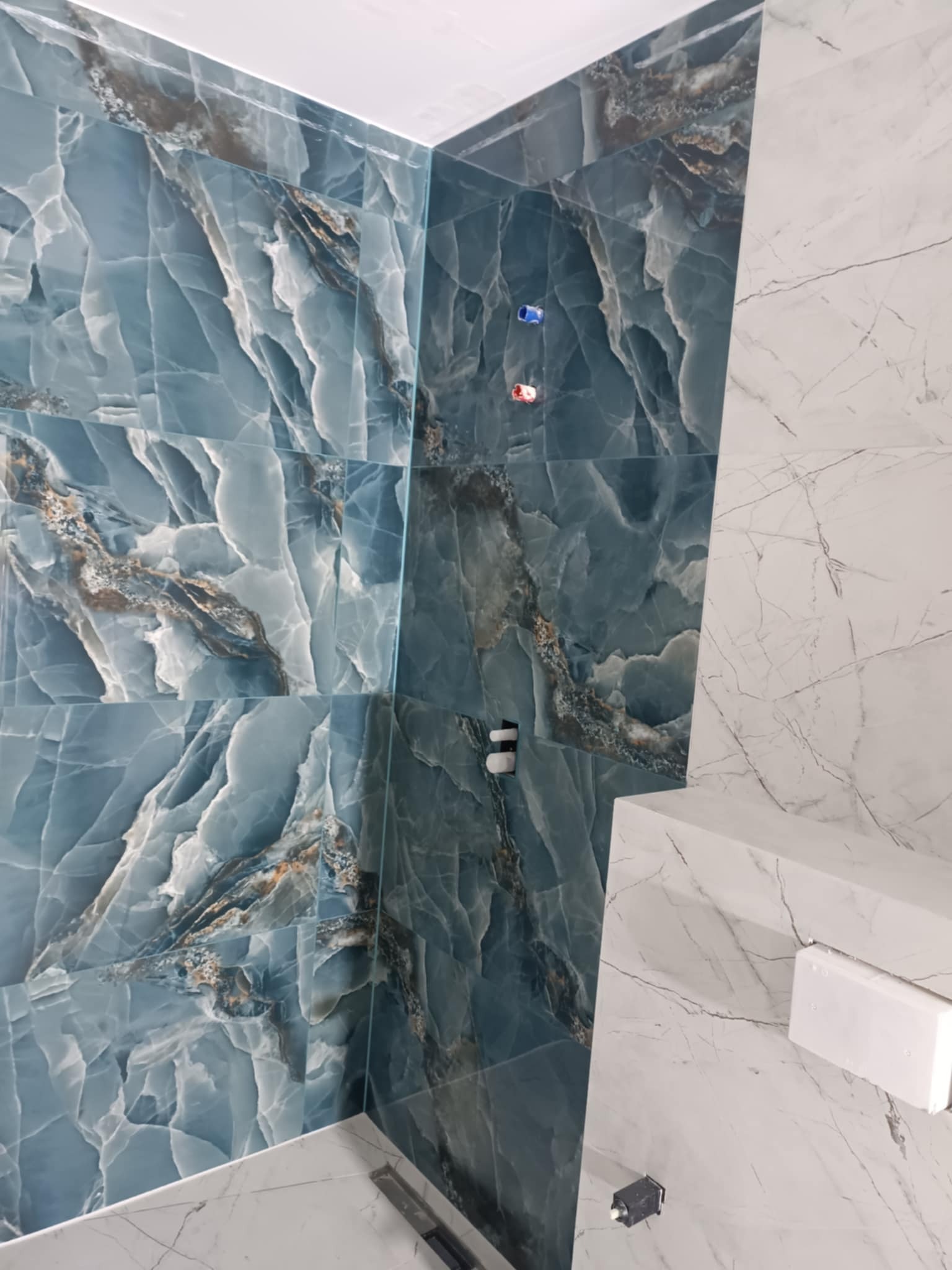 Ściana łazienki wyłożona niebieskimi płytkami imitującymi onyks, kontrastująca z białymi płytkami marmuropodobnymi na sąsiedniej ścianie, widoczne przyłącza wodne.