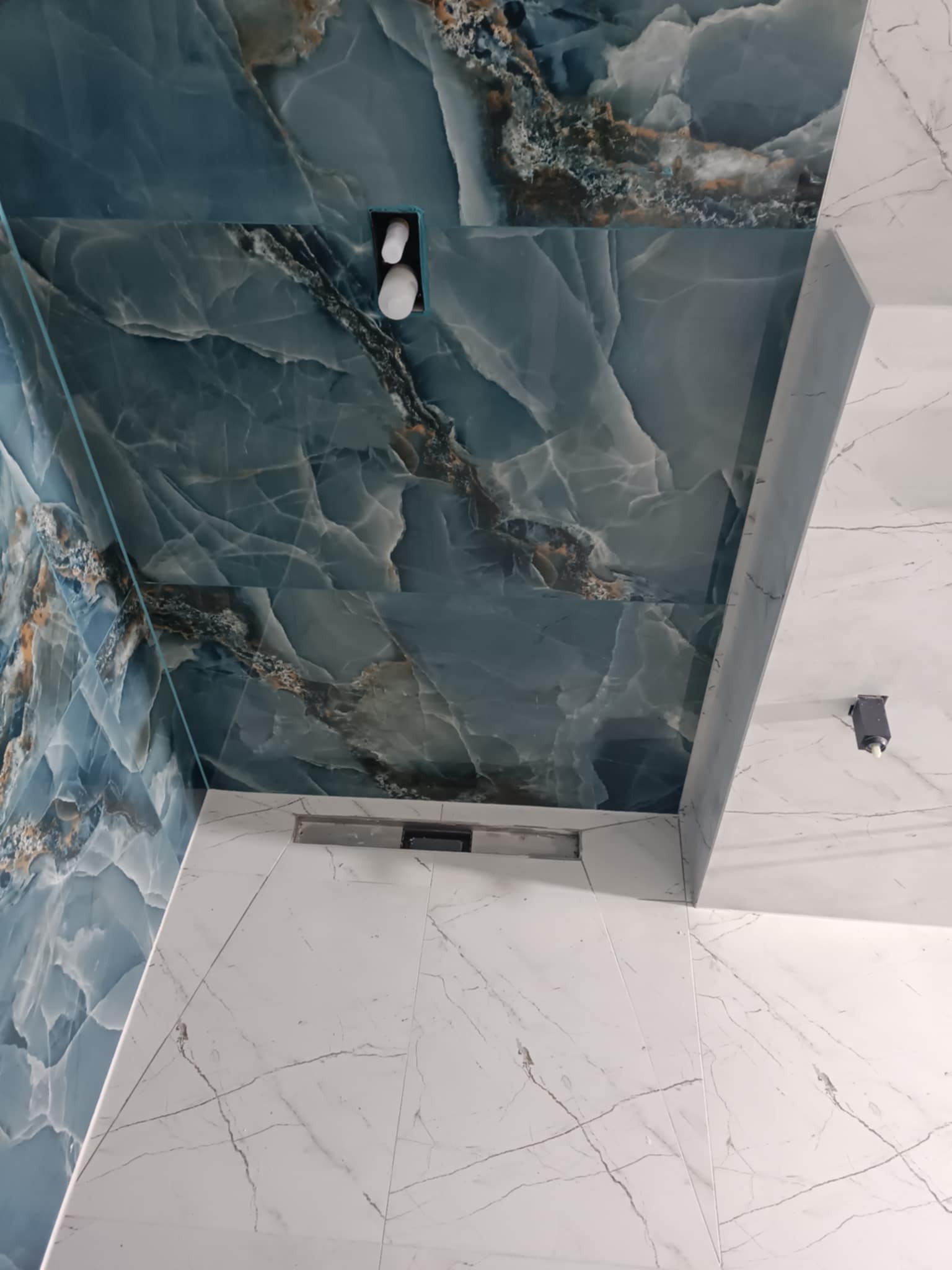 Wykończona łazienka z niebieskimi płytkami imitującymi marmur na ścianie i suficie, oraz białymi płytkami podłogowymi z szarymi żyłkami, widoczny odpływ liniowy i punktowe oświetlenie.