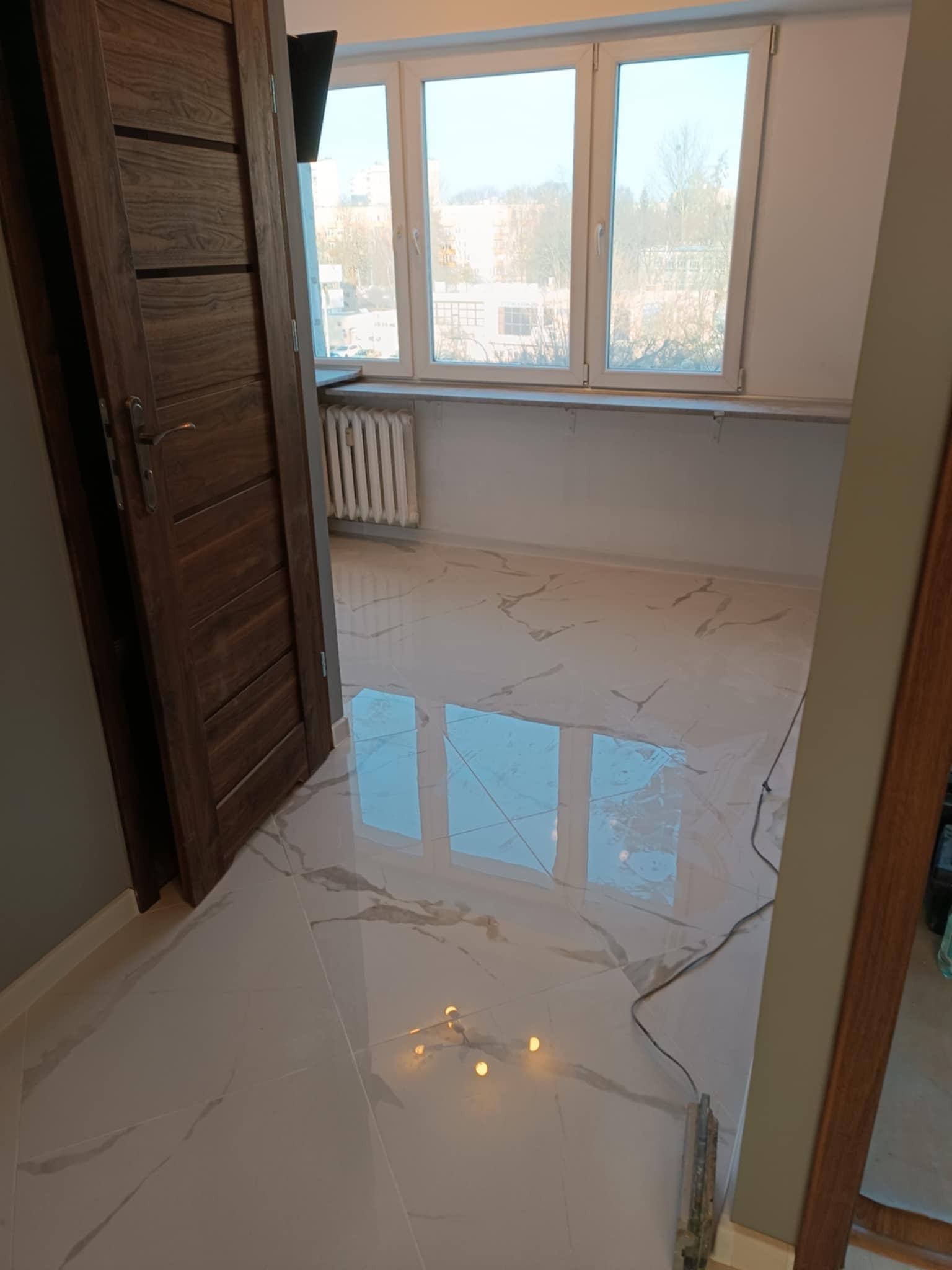 Nowo ułożona, błyszcząca podłoga z płytek imitujących marmur w jasnym pomieszczeniu z oknem i grzejnikiem. Widoczna otwarta, drewniana rama drzwi i narzędzia do układania płytek przy podłodze.