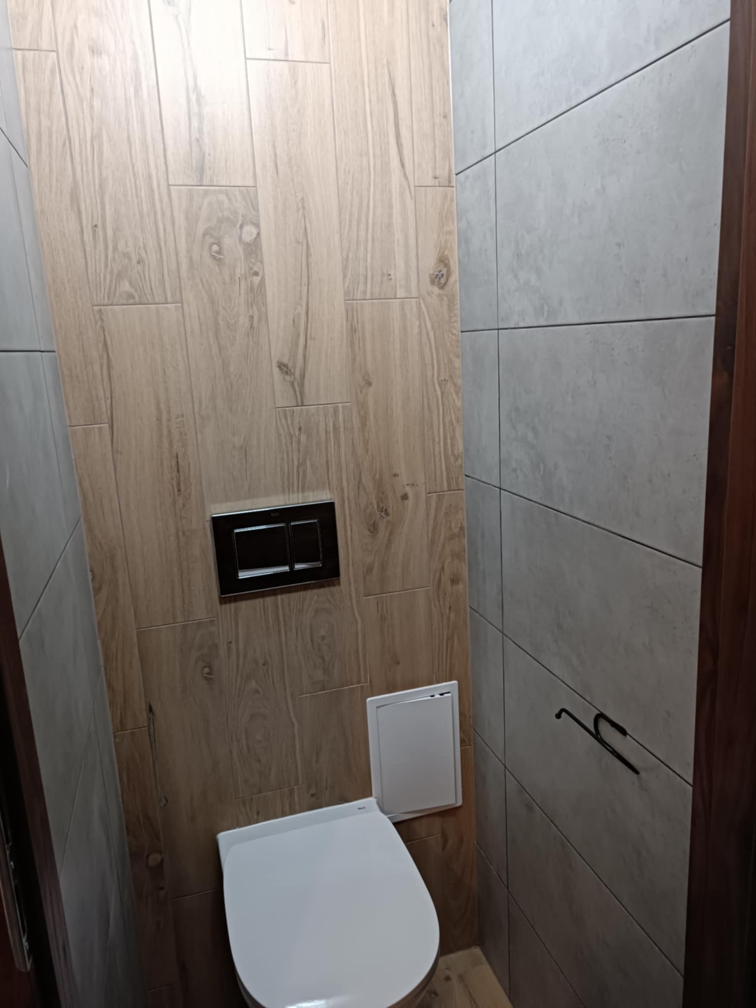 Nowoczesna toaleta podwieszana z ukrytą spłuczką, obudowana płytkami imitującymi drewno i szare płytki, z czarnym przyciskiem spłukującym i metalowym wieszakiem na ręczniki.