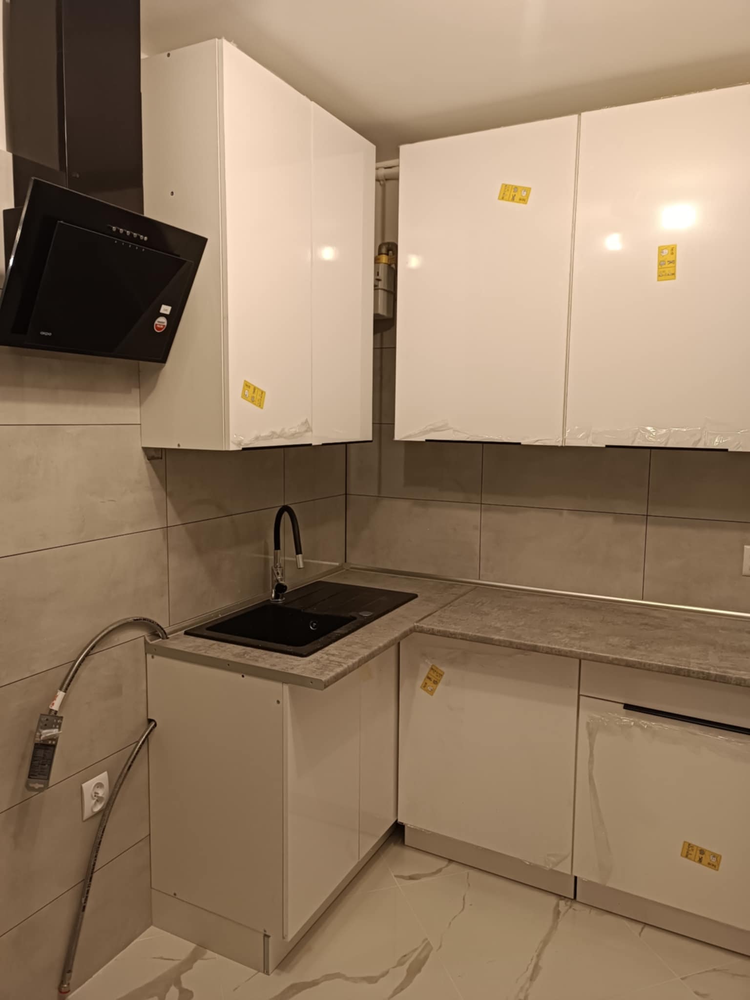 Nowa, jasna kuchnia z czarnym zlewem i okapem, jeszcze z folią ochronną na frontach szafek, płytki imitujące kamień na ścianie.