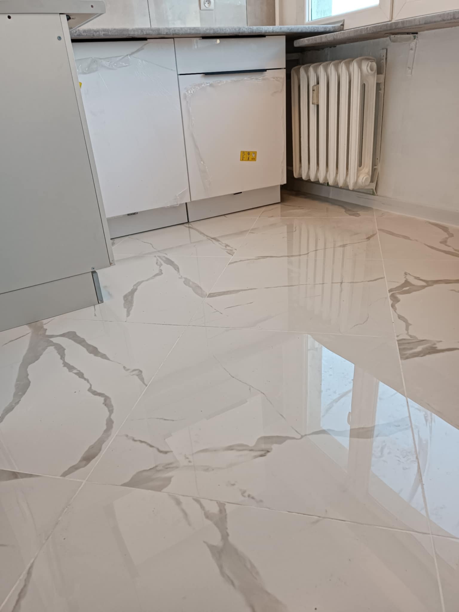 Błyszczące, białe płytki podłogowe imitujące marmur, ułożone w nowym pomieszczeniu z grzejnikiem i częściowo widocznymi białymi szafkami kuchennymi z folią ochronną.