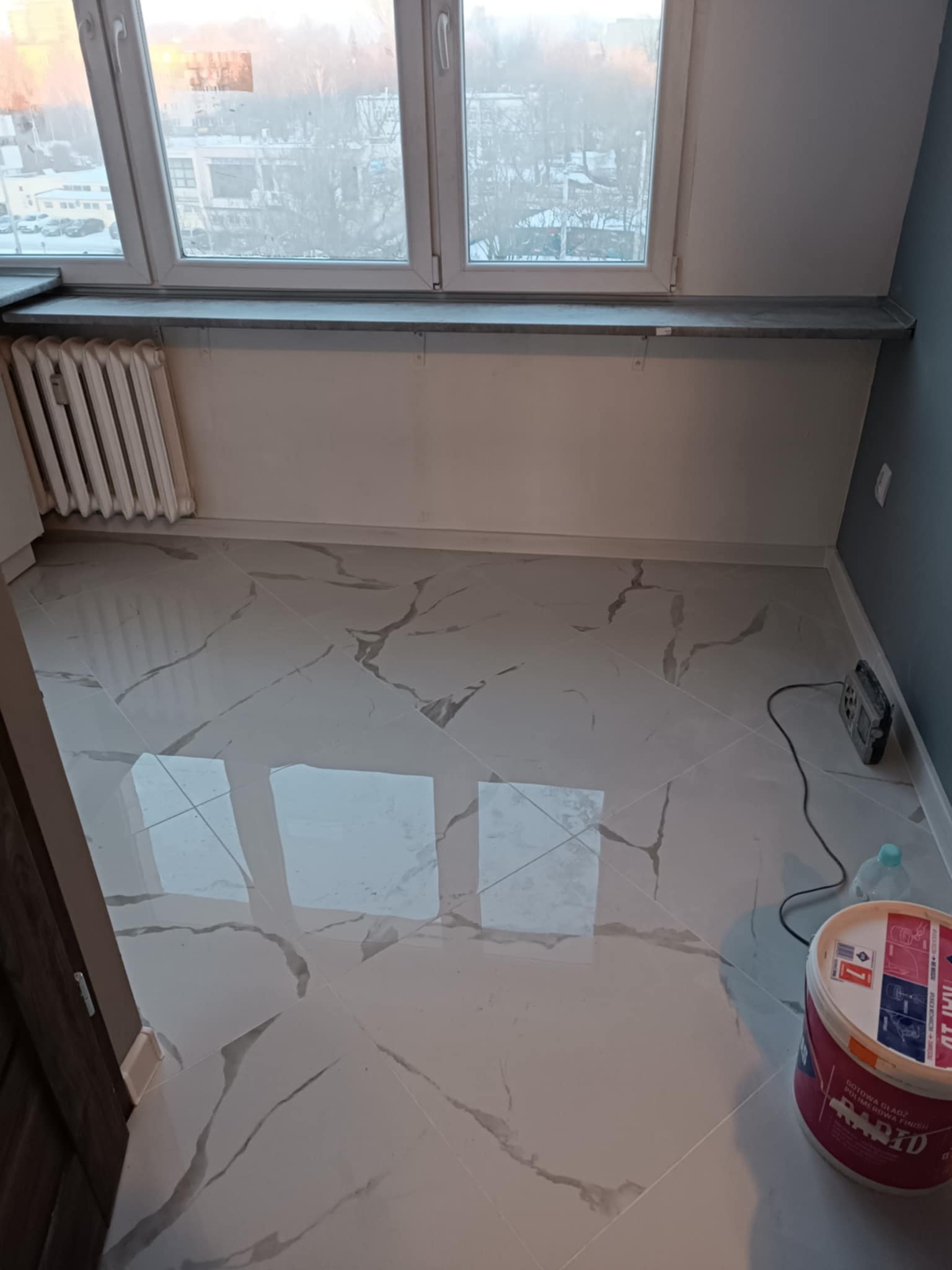 Świeżo położone, błyszczące płytki podłogowe imitujące marmur w pokoju z grzejnikiem pod oknem, widoczny pojemnik z gotową gładzią polimerową oraz radio budowlane.