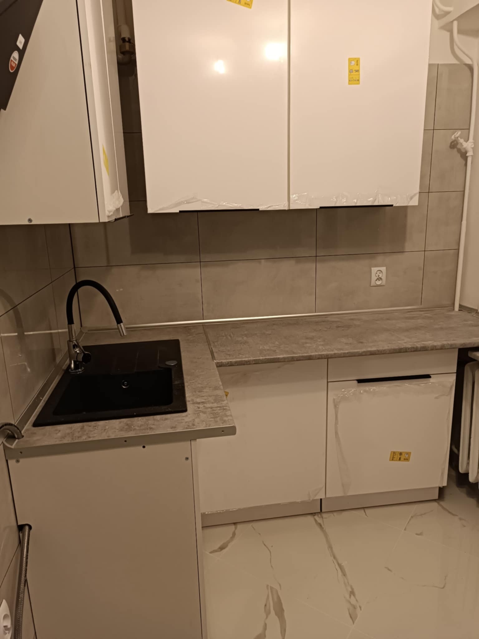 Nowoczesna, minimalistyczna kuchnia w trakcie wykańczania, z białymi szafkami z folią ochronną, czarnym zlewem z elastyczną wylewką i szarymi płytkami na ścianie, widoczne gniazdko elektryczne.