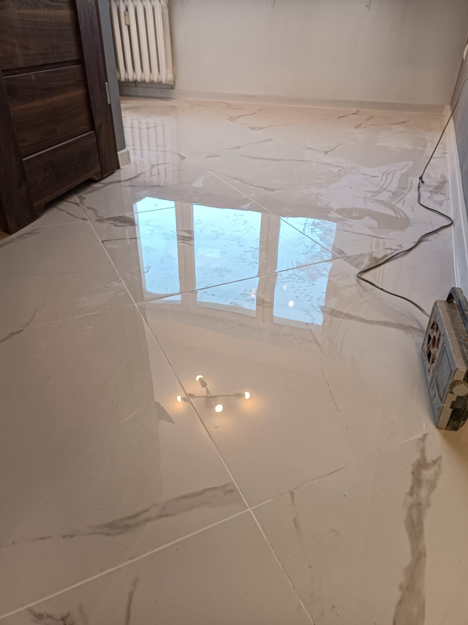 Jasne, polerowane płytki podłogowe z szarymi żyłkami, odbijające okno, z widocznym grzejnikiem, szafką i kablem z urządzeniem na podłodze.