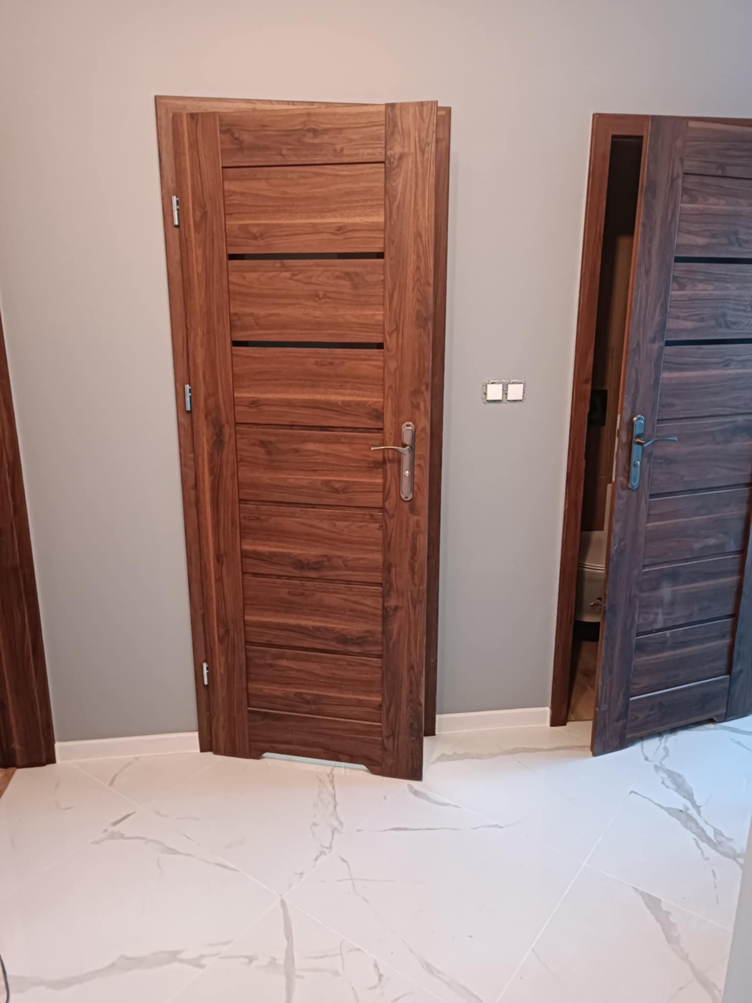 Dwa drewniane drzwi wewnętrzne z poziomymi wstawkami, jeden zamknięty, drugi otwarty, na tle szarej ściany i białej podłogi z marmurowym wzorem.
