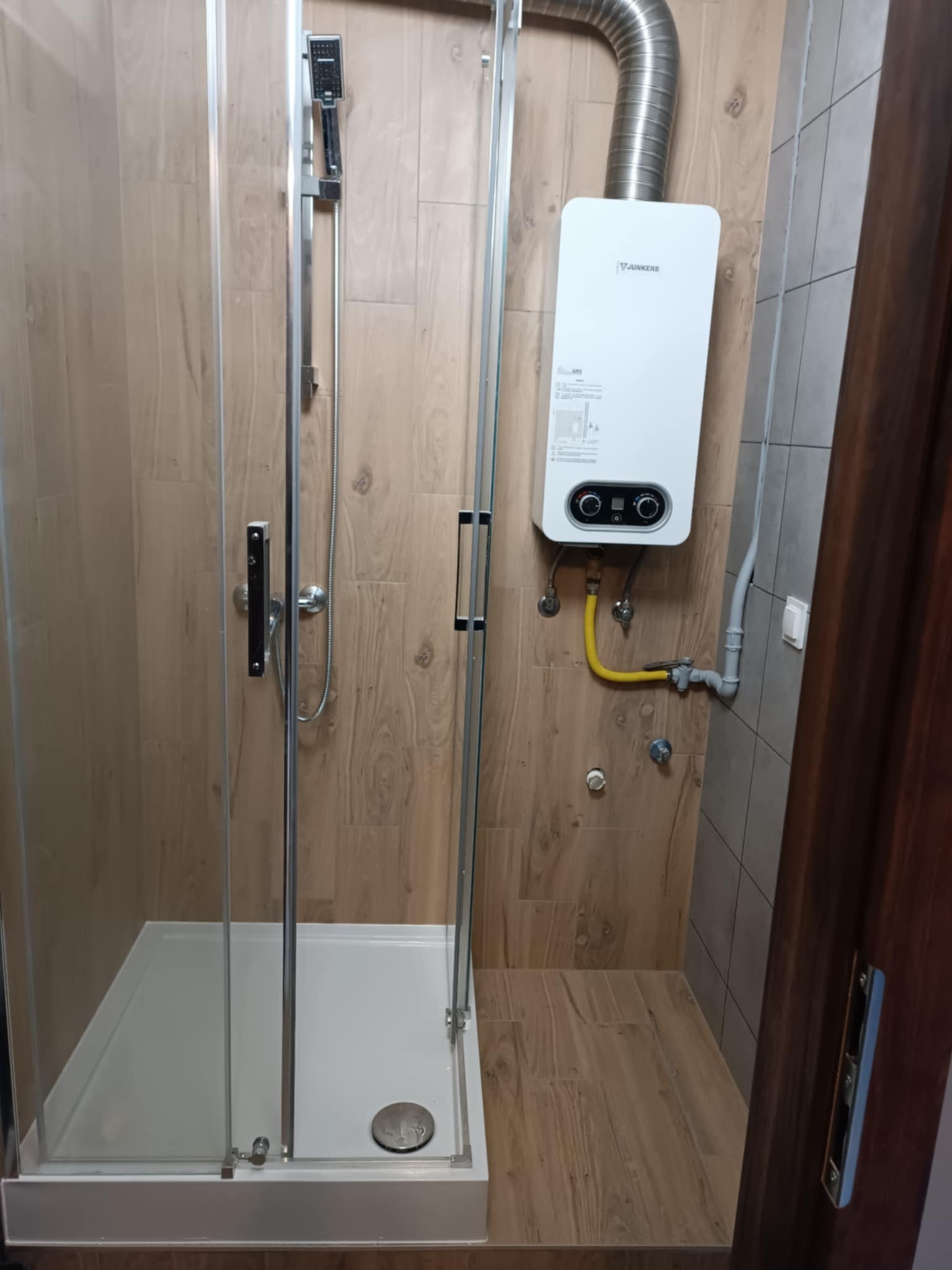 Kabina prysznicowa z zamontowanym piecem gazowym Junkers na ścianie wyłożonej płytkami imitującymi drewno, z żółtą instalacją gazową.