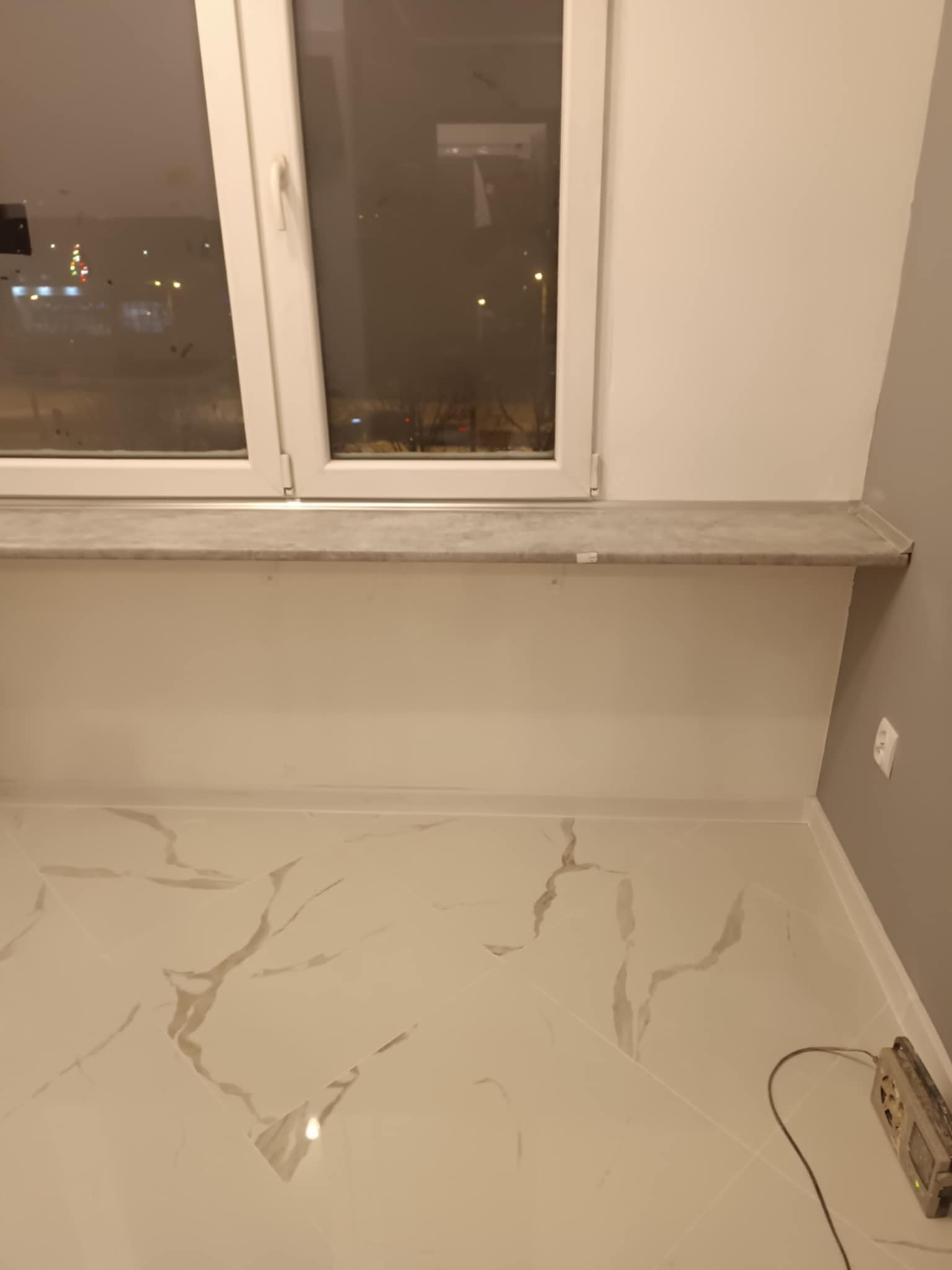 Wykończona wnęka okienna z parapetem z płytek imitujących marmur, białe ściany i podłoga z płytek w podobnym stylu, widoczny fragment szarej ściany oraz narzędzie pomiarowe przy podłodze.