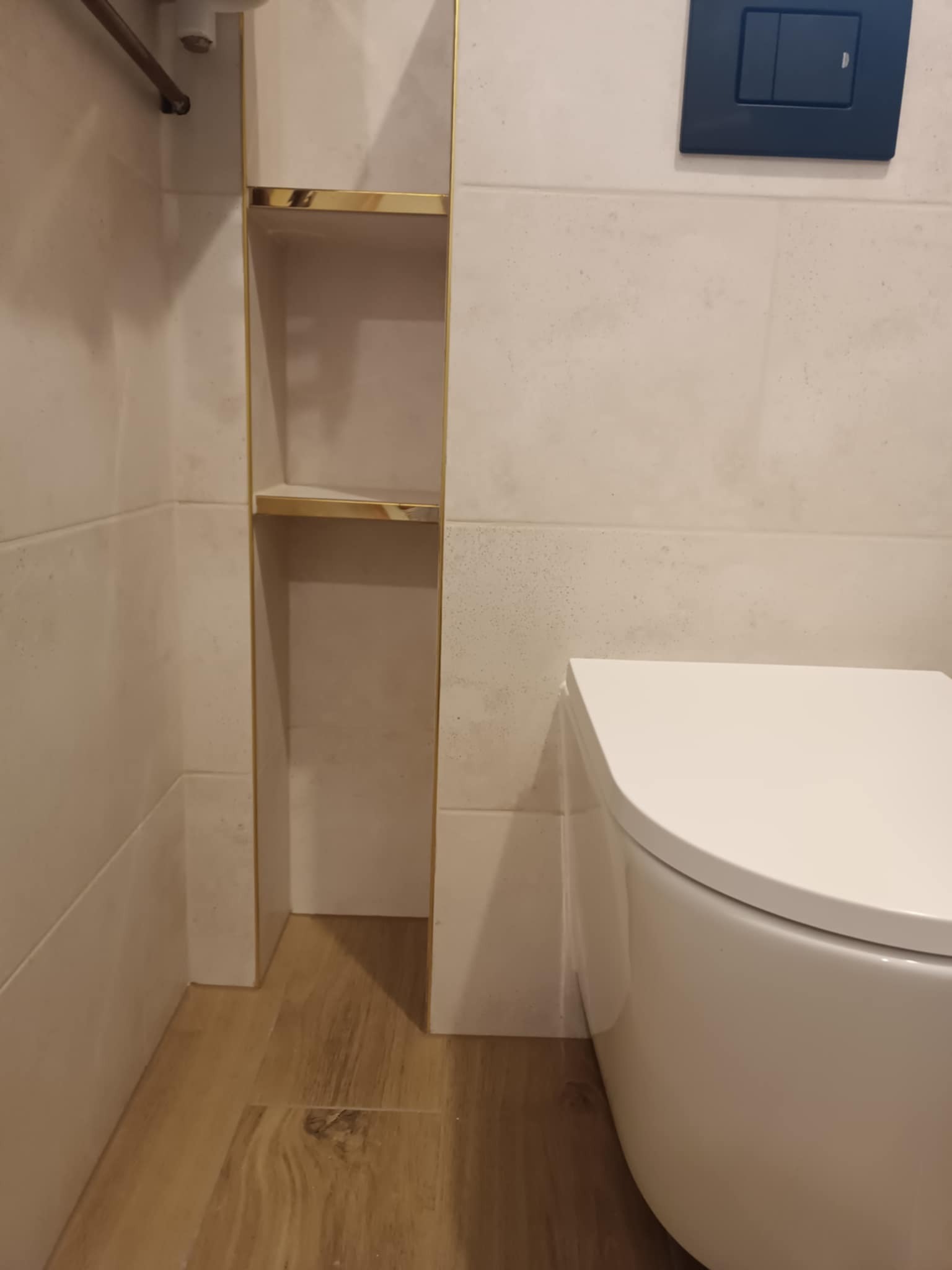 Nowoczesna toaleta wisząca obok wnęki z półkami wykończonej złotymi listwami, na ścianie kremowe płytki imitujące kamień i czarny przycisk spłuczki.