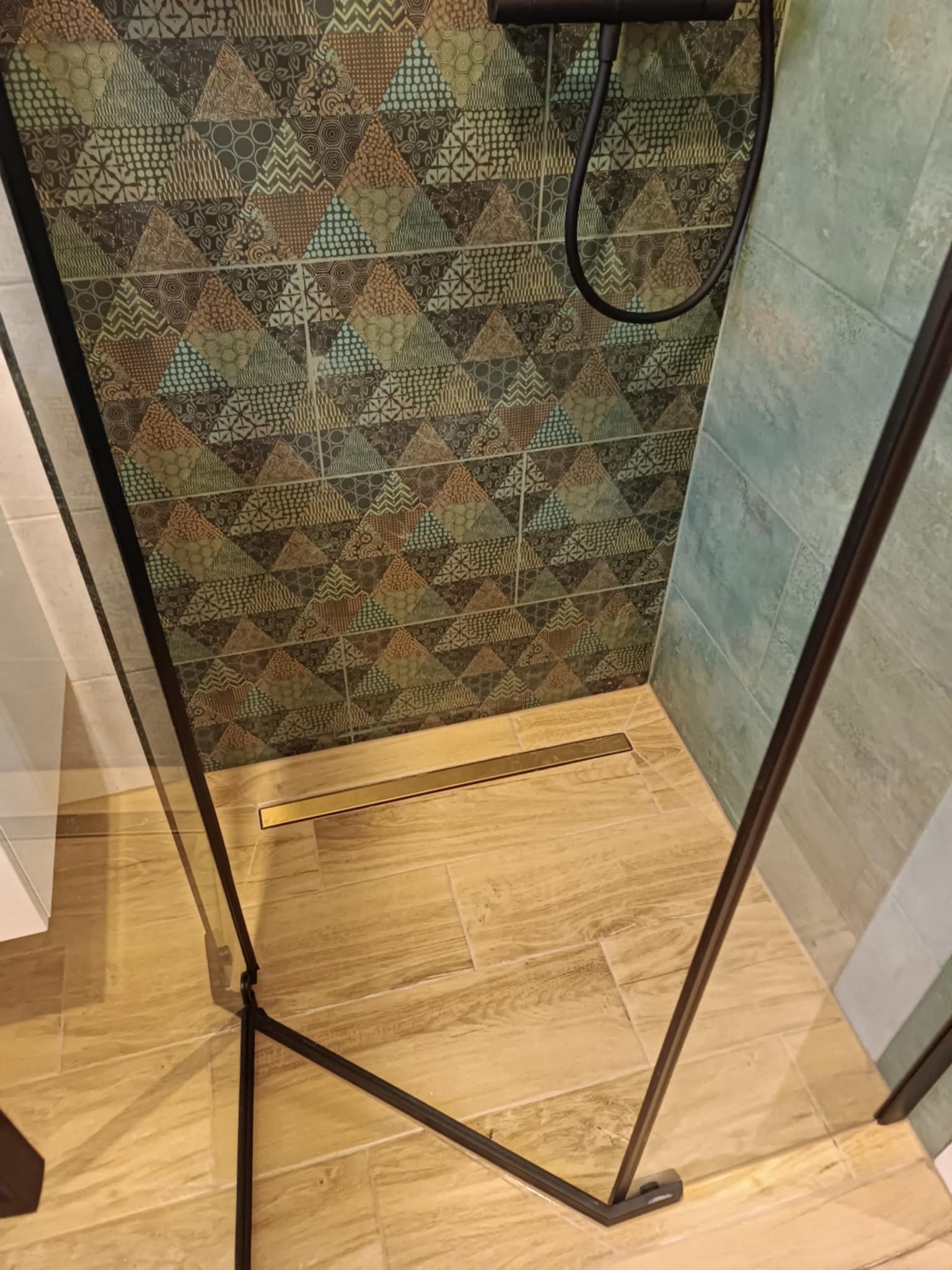 Kabina prysznicowa z czarnymi profilami, wyłożona płytkami o zróżnicowanych wzorach geometrycznych i kolorach, z odpływem liniowym w podłodze.