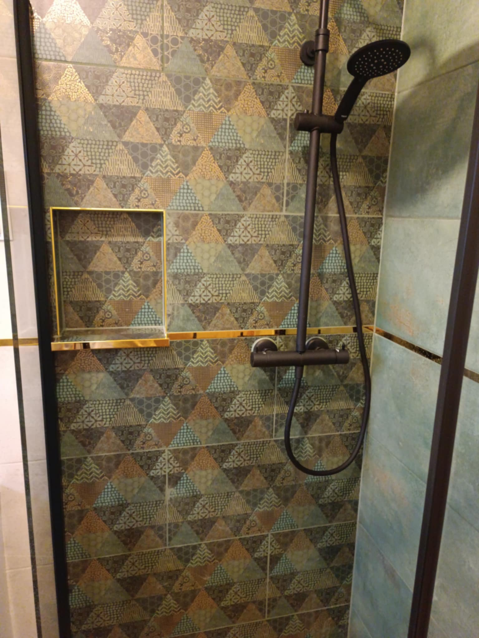 Kabina prysznicowa z geometrycznymi płytkami w odcieniach brązu, złota i turkusu, wyposażona w czarną armaturę i wnękę z półką wykończoną złotą listwą.