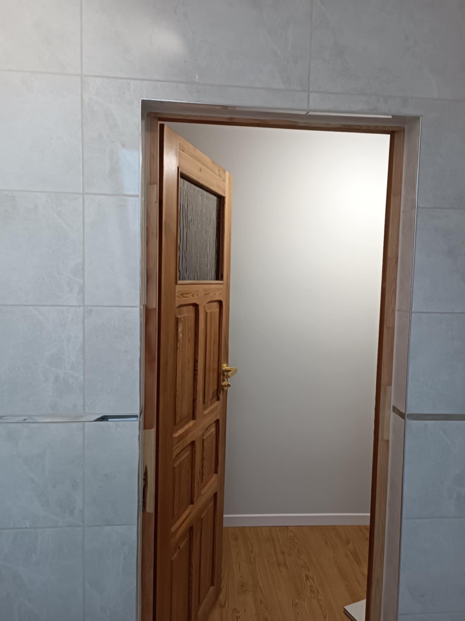 Otwarta drewniana drzwi z szybą w łazience z jasnymi, marmurkowymi płytkami na ścianach i panelami podłogowymi wewnątrz.