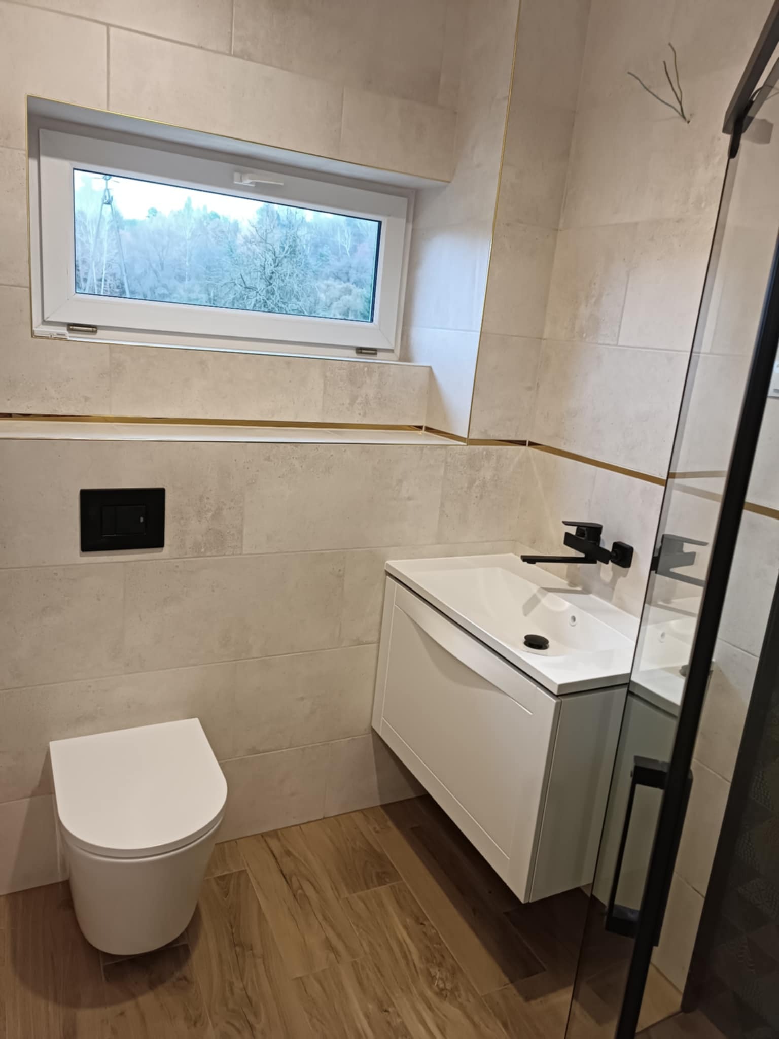 Nowoczesna łazienka z beżowymi płytkami, podwieszaną toaletą, białą szafką z umywalką i czarną armaturą, widok na zewnątrz przez okno.