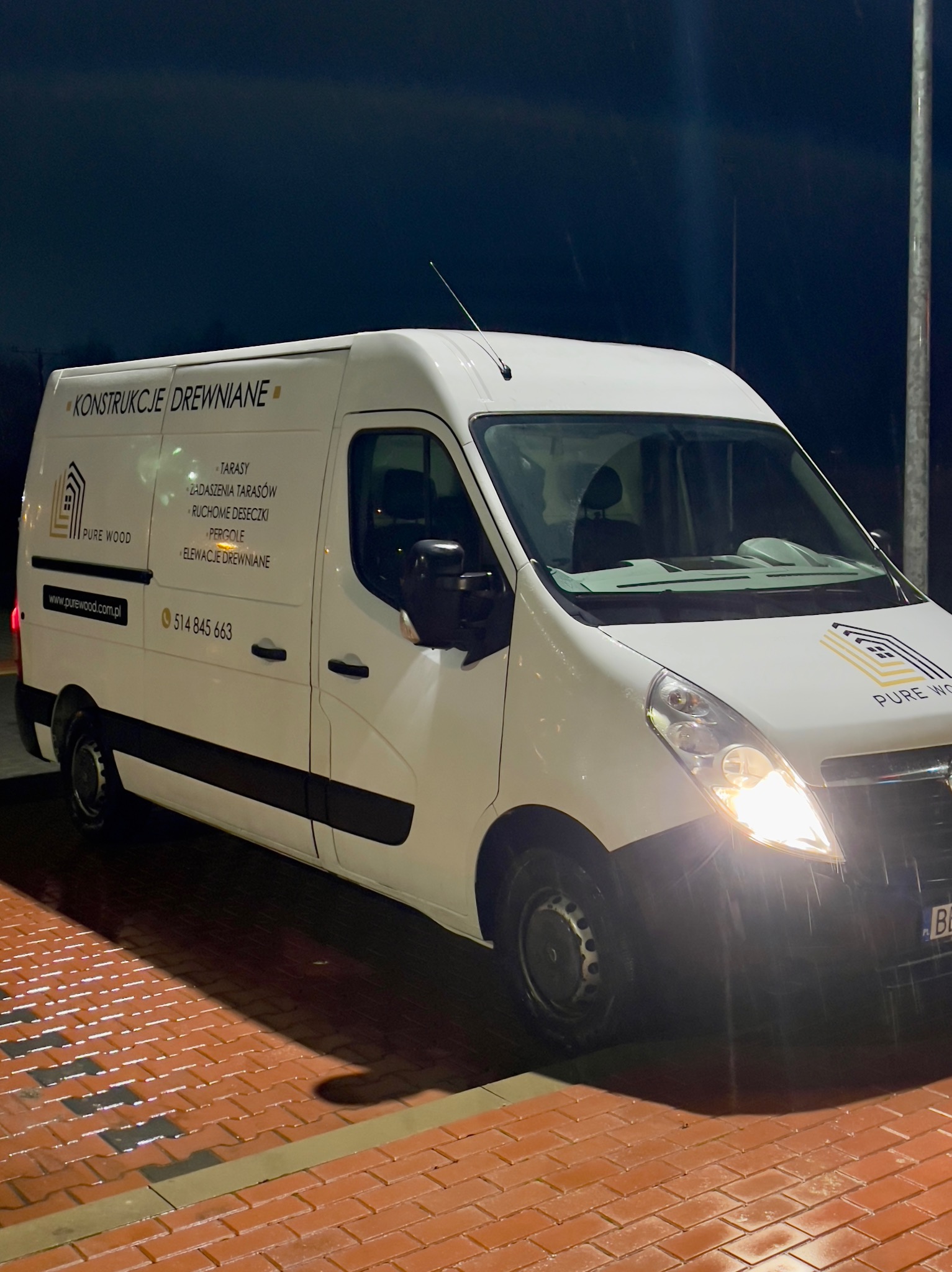 Biały samochód dostawczy z logo i informacjami o firmie Pure Wood, specjalizującej się w konstrukcjach drewnianych, tarasach i elewacjach. Widoczne oświetlenie i mokra nawierzchnia.