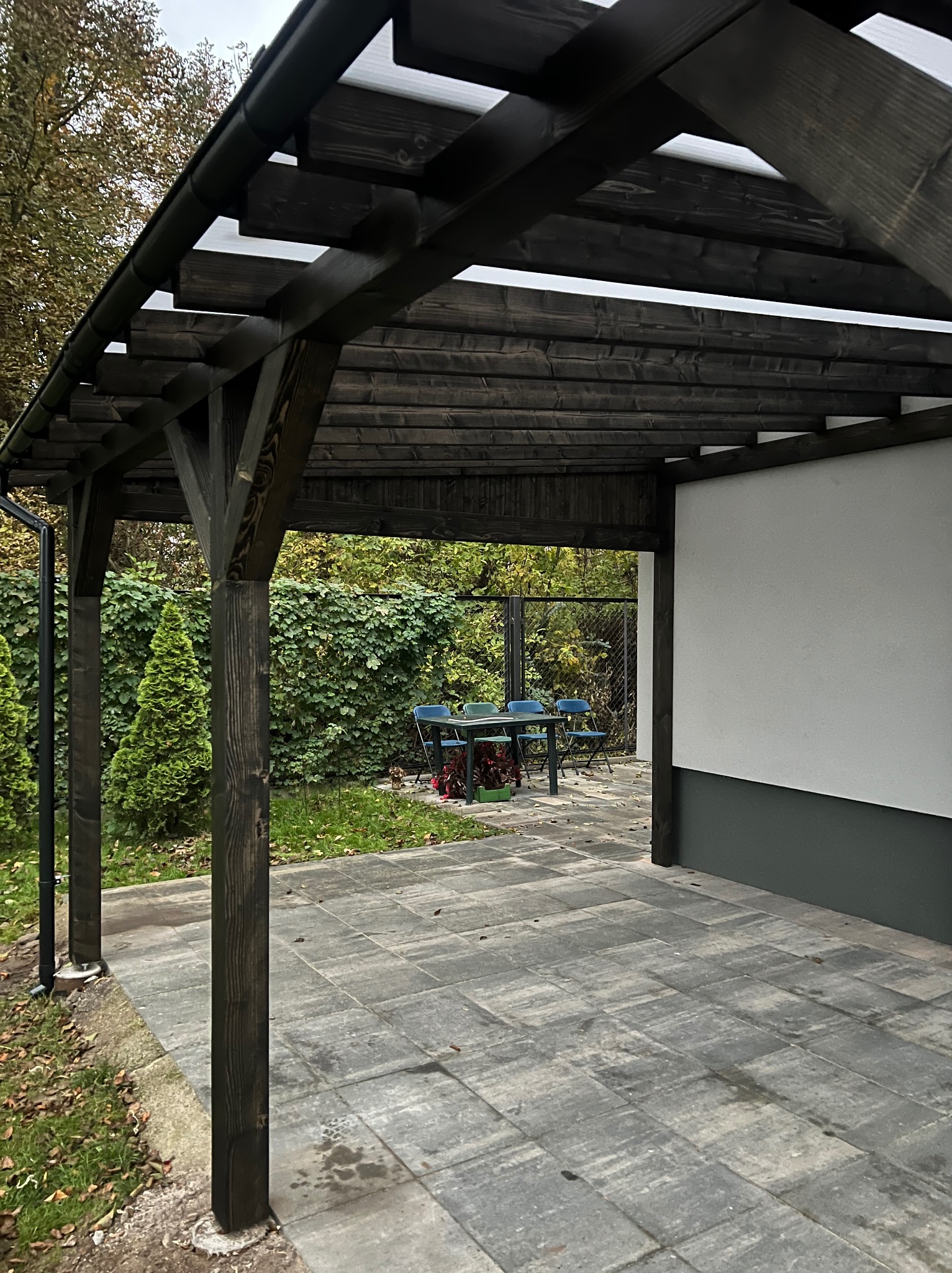 Ciemna, drewniana pergola z przezroczystym zadaszeniem, z widocznym stołem i krzesłami w tle, na tle zieleni. Ułożona kostka brukowa na podłożu.