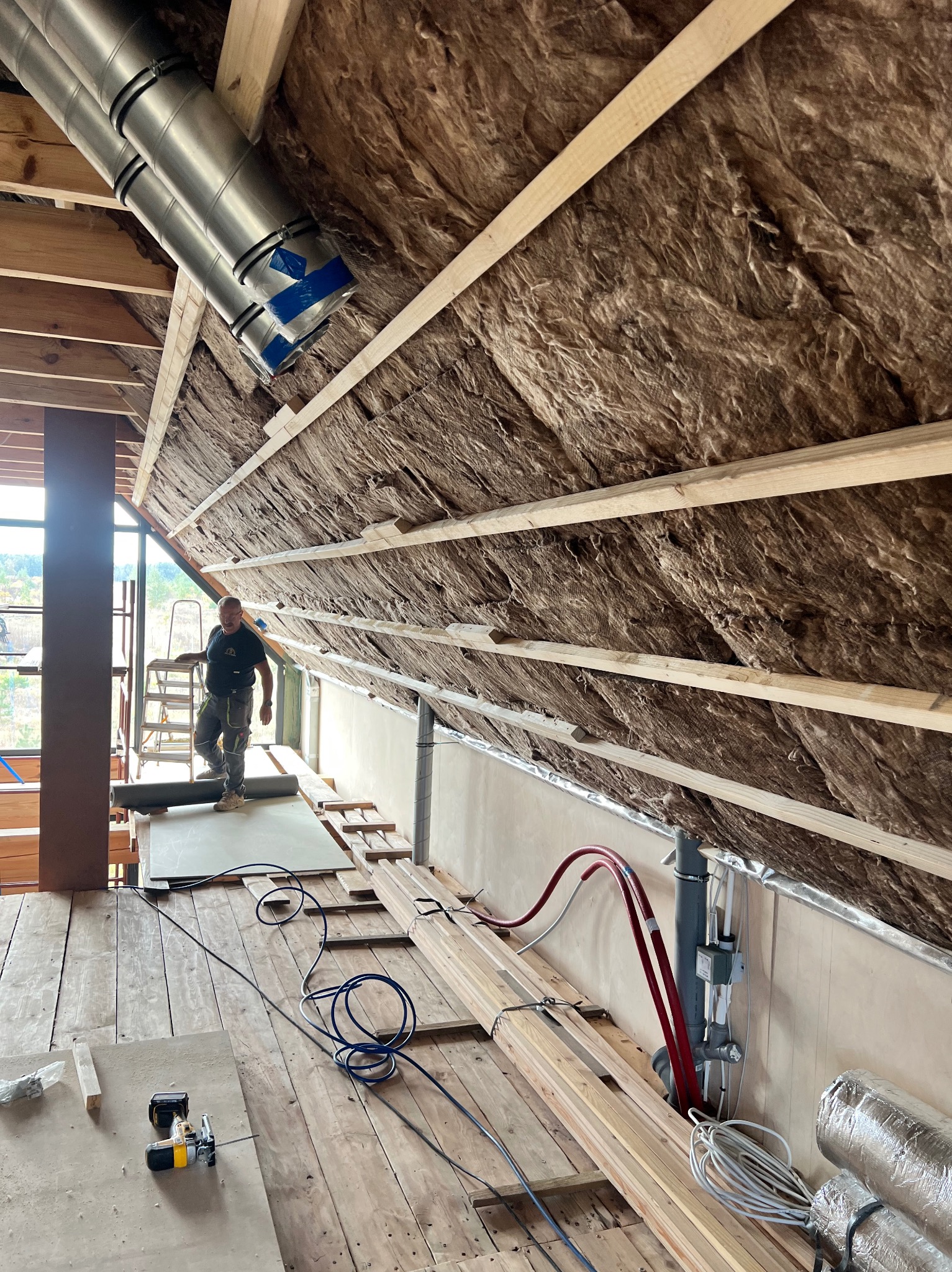 Montaż izolacji poddasza z widocznymi elementami konstrukcyjnymi, instalacją wentylacyjną i pracownikiem na drabinie. Widoczne narzędzia i materiały budowlane.