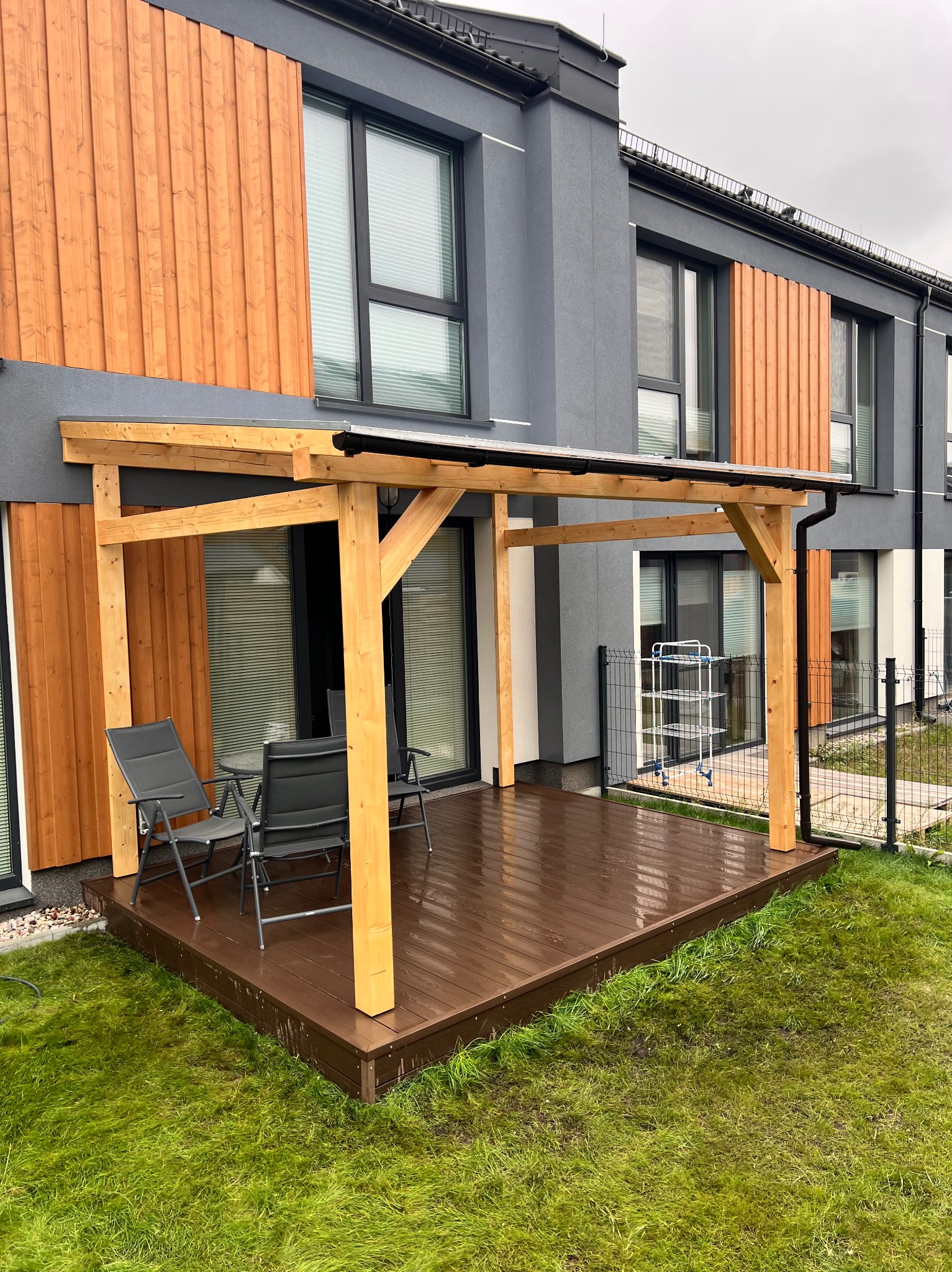 Nowoczesna pergola z jasnego drewna na brązowym tarasie, z szarymi krzesłami i stolikiem. Tło stanowi dwukondygnacyjny dom z elementami drewnianymi.