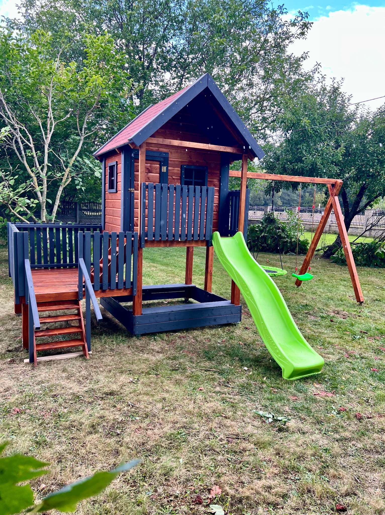 Drewniany plac zabaw z domkiem, zjeżdżalnią i huśtawką na trawniku. Elementy pomalowane na brązowo i grafitowo. Widok z bliska, w tle drzewa i ogrodzenie.