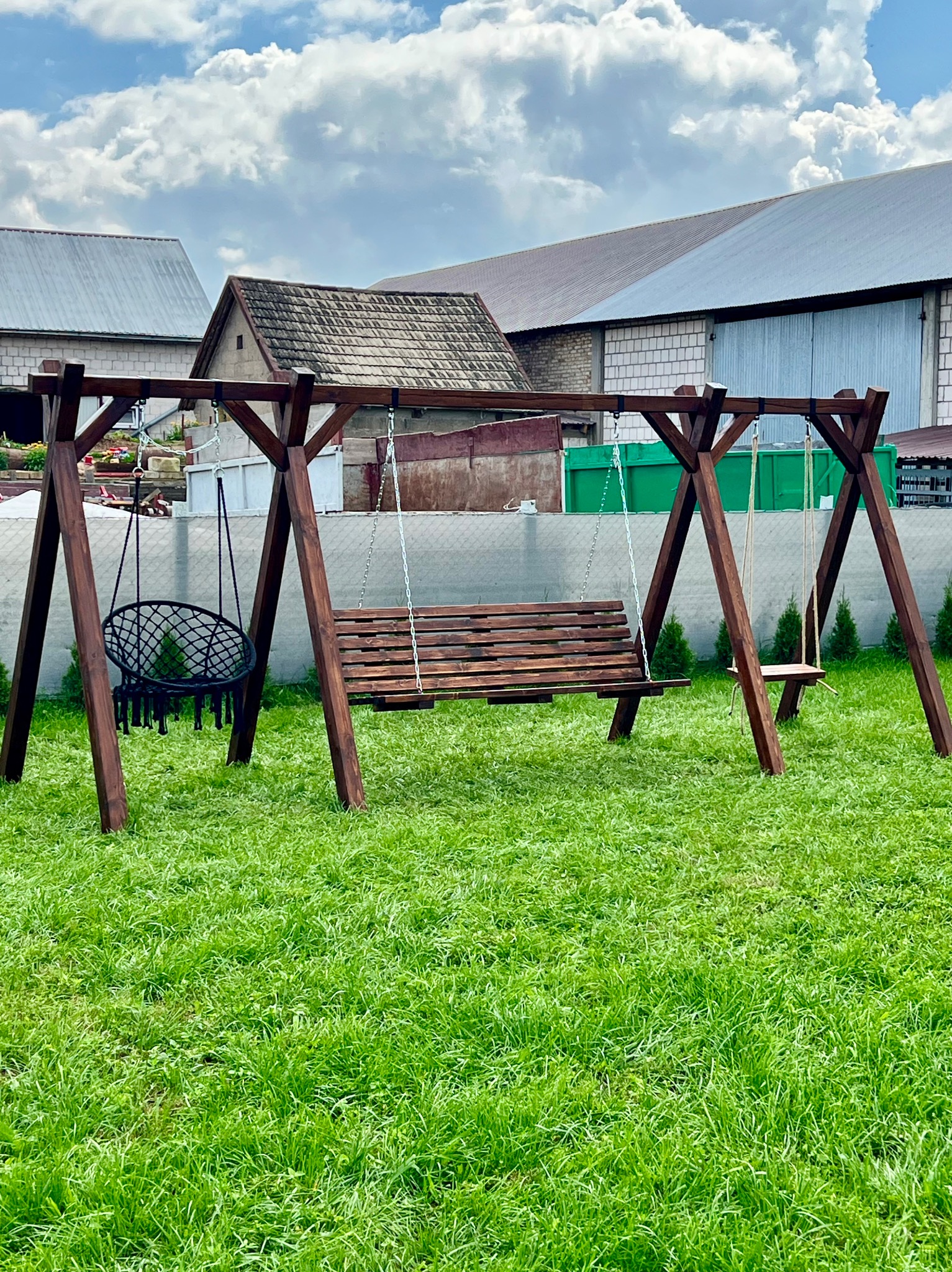 Drewniana huśtawka ogrodowa z trzema siedziskami: ławka, pojedyncze siedzisko i okrągłe krzesło wiszące, na tle zielonego trawnika i budynków gospodarczych.