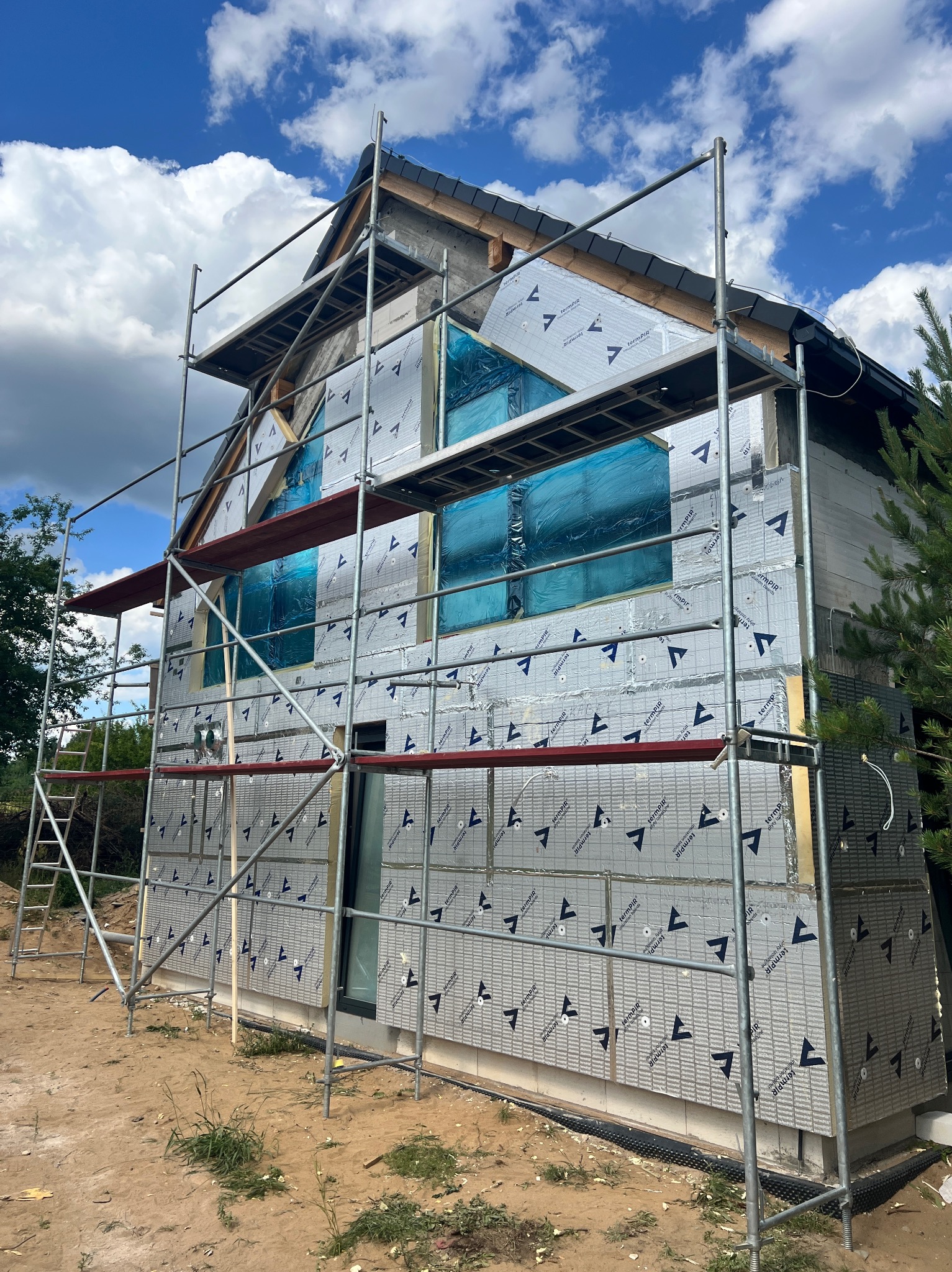 Budowa domu jednorodzinnego w trakcie ocieplania, widoczne rusztowania i materiały izolacyjne z nadrukiem, okna zabezpieczone folią. Słoneczny dzień.