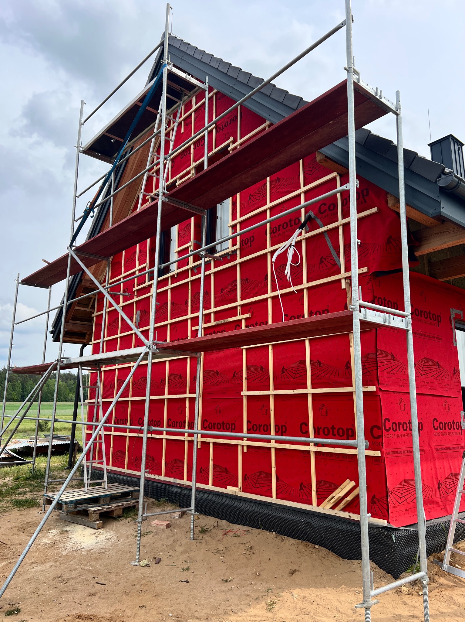 Dom w trakcie ocieplania, widoczne rusztowania, czerwona membrana i drewniane łaty. Prace budowlane w toku, widok z zewnątrz na elewację domu.