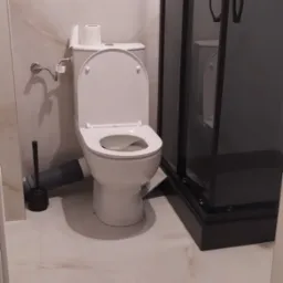 Biała toaleta z otwartą klapą i widoczną rurą odpływową, obok czarna szczotka do WC i fragment kabiny prysznicowej z czarną ramą, na podłodze płytki imitujące marmur.