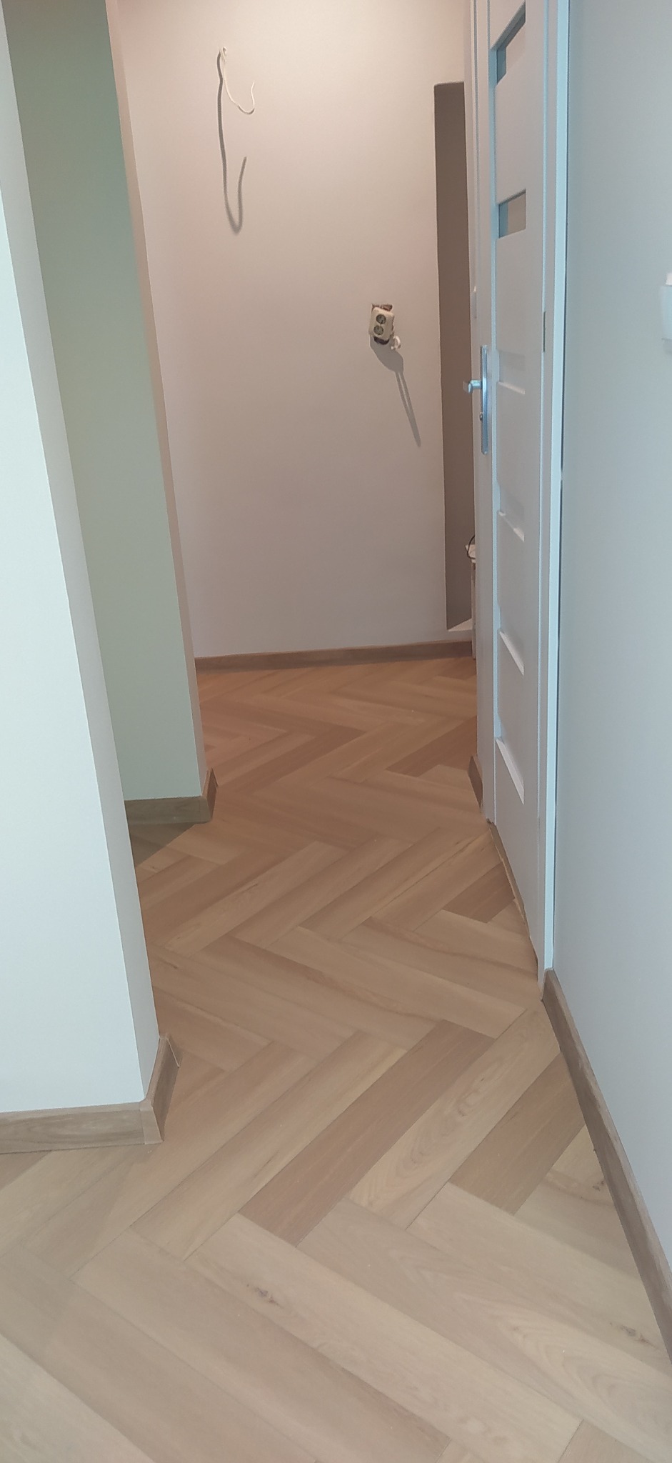Ułożony parkiet w jodełkę w jasnym korytarzu, widoczne niedokończone wykończenia ścian i gniazdko elektryczne.