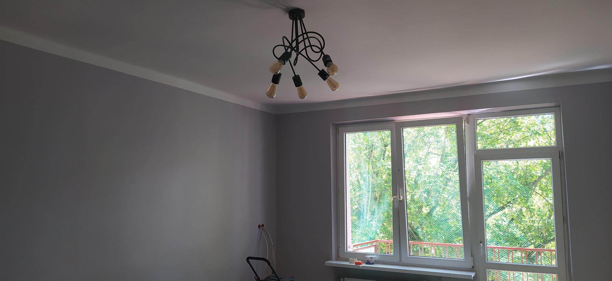 Pokój po malowaniu ścian na szaro, widoczny dekoracyjny sufitowy żyrandol z żarówkami Edisona, biała listwa przysufitowa i okno z widokiem na zieleń.