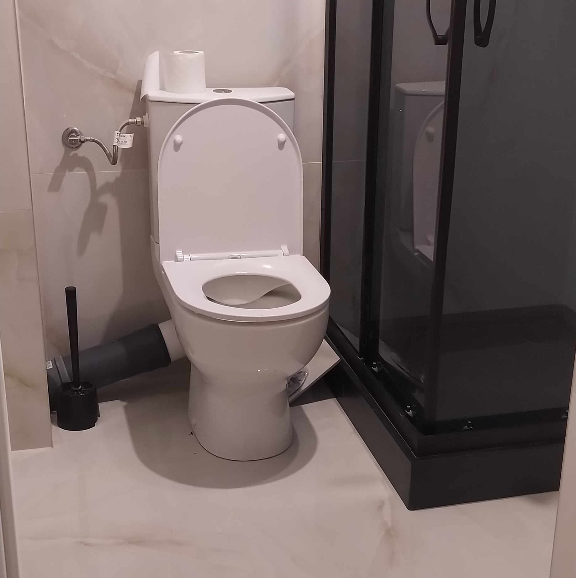 Biała toaleta z otwartą klapą i widoczną rurą odpływową, obok czarna szczotka do WC i fragment kabiny prysznicowej z czarną ramą, na podłodze płytki imitujące marmur.
