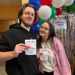 Rozbawieni Events - Dwoje uśmiechniętych animatorów z ulotkami 'Rozbawieni Events' i lizakami na tle dekoracji z balonów i błyszczącej kurtyny. #SportoweSerce