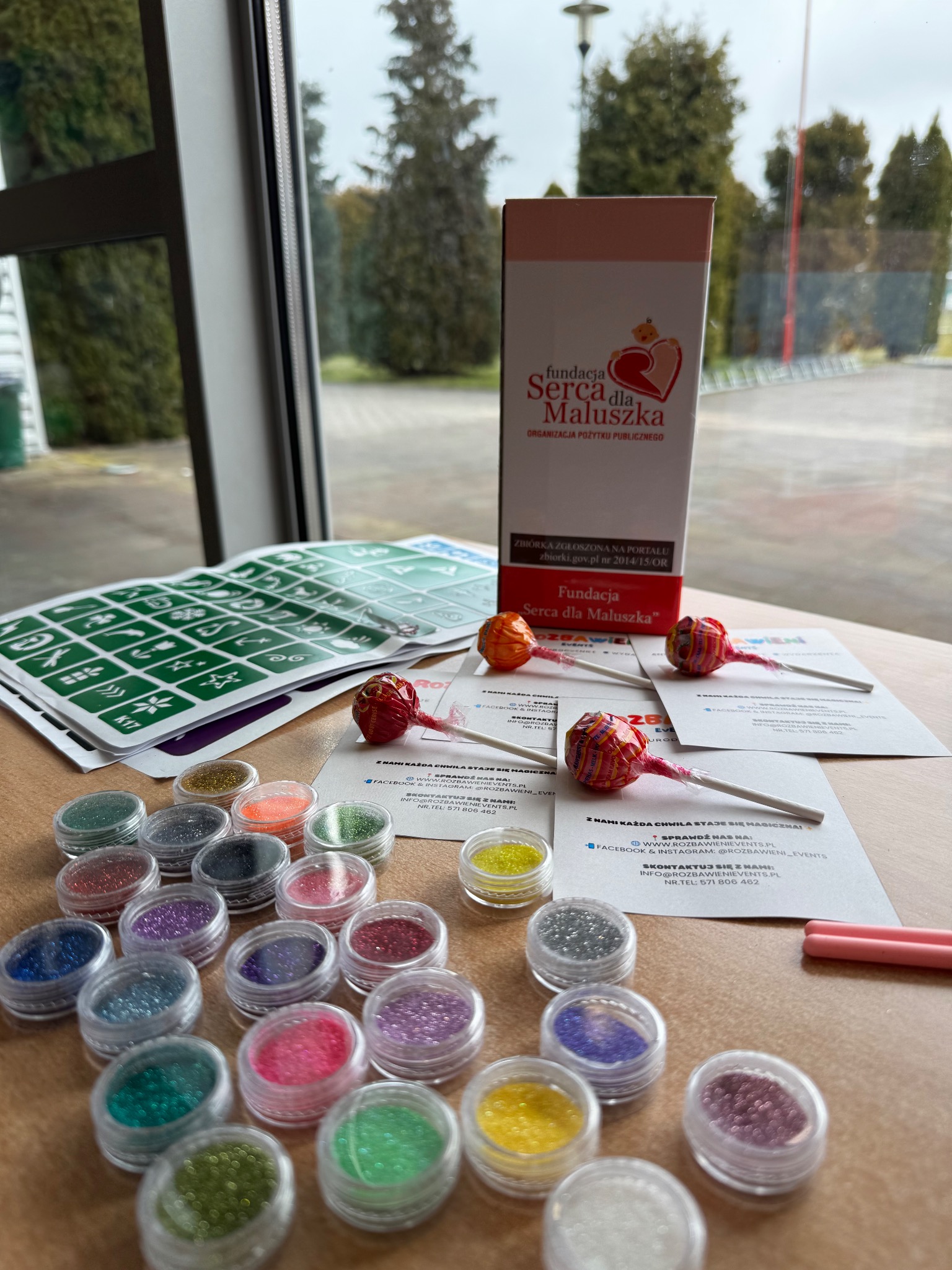 Akcesoria do malowania twarzy: szablony, brokaty, lizaki i wizytówki firmy eventowej na stole przy oknie z widokiem na zewnątrz, w neutralnym oświetleniu.