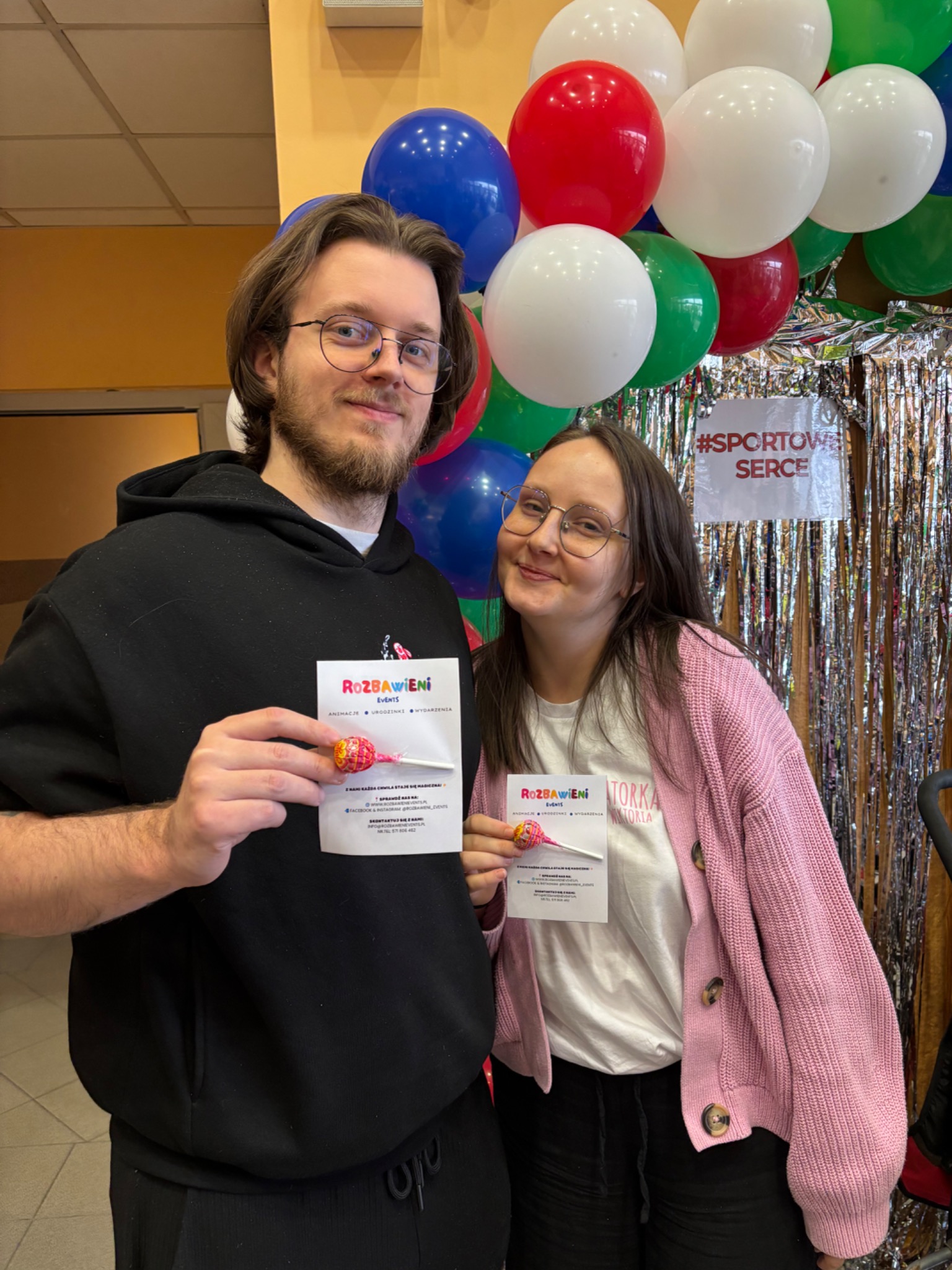 Dwoje uśmiechniętych animatorów z ulotkami 'Rozbawieni Events' i lizakami na tle dekoracji z balonów i błyszczącej kurtyny. #SportoweSerce
