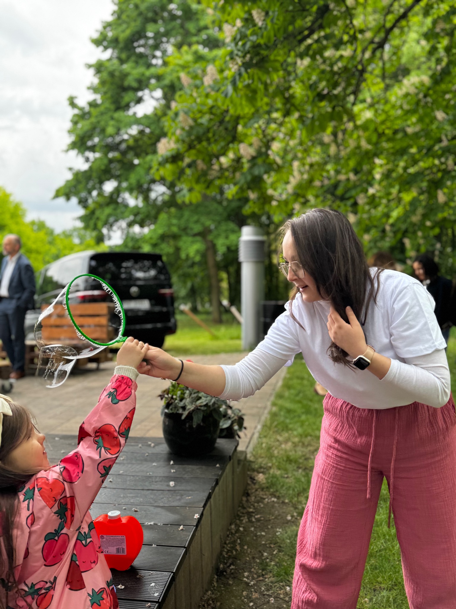Animator puszcza bańki mydlane dziewczynce w różowej kurtce w truskawki. W tle zieleń drzew, mężczyzna w garniturze i czarny van.