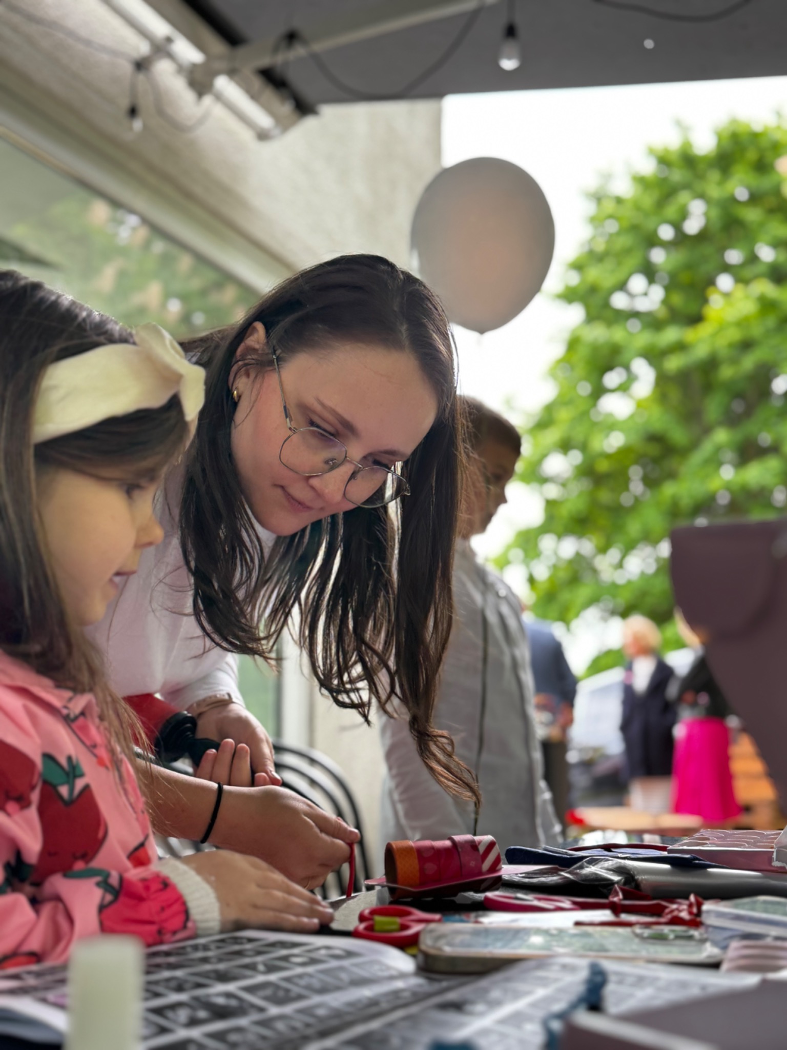 Animator pomaga dziewczynce w warsztatach rękodzielniczych na świeżym powietrzu. Na stole materiały do robótek. W tle balon i zieleń.