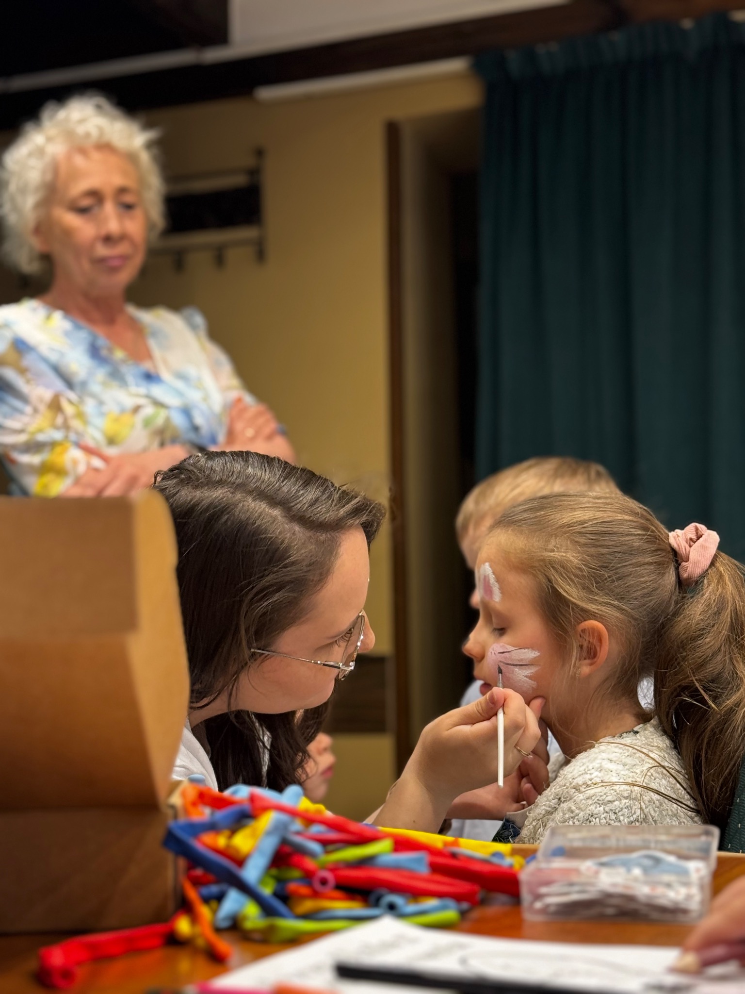 Animator maluje twarz dziewczynki pędzlem. W tle stoi starsza kobieta z założonymi rękami. Na stole balony i akcesoria do malowania.