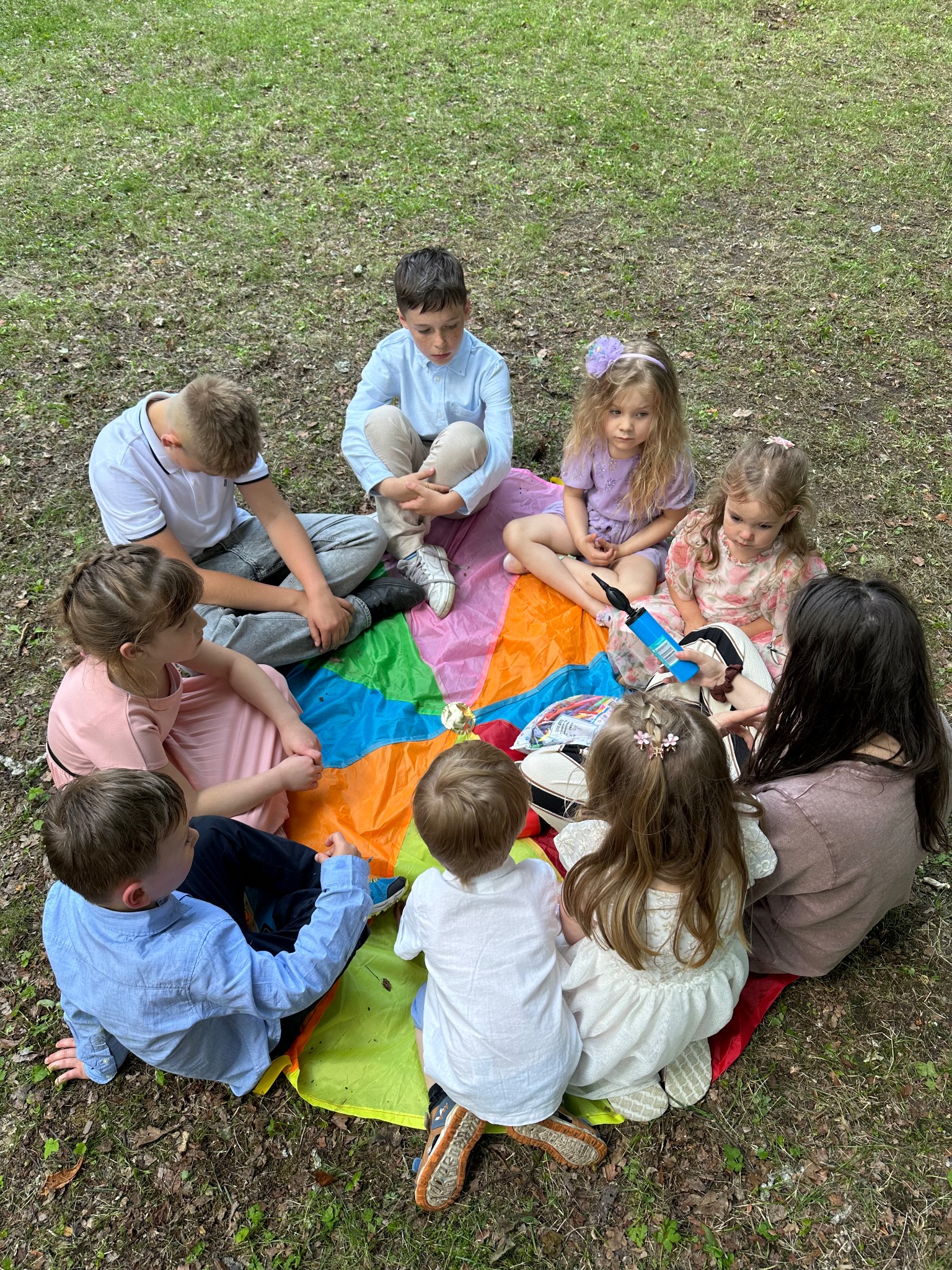 Grupa dzieci siedzi na kolorowej chuście na trawie, otoczona przez dorosłego. Zabawa na świeżym powietrzu w parku, z bliska widoczna pompka do balonów.