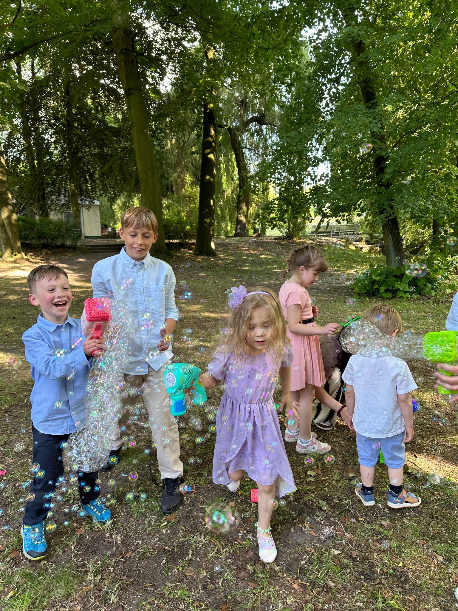 Grupa dzieci bawi się na trawie, puszczając bańki mydlane z kolorowych pistoletów, w tle drzewa i budynek, radosna atmosfera.