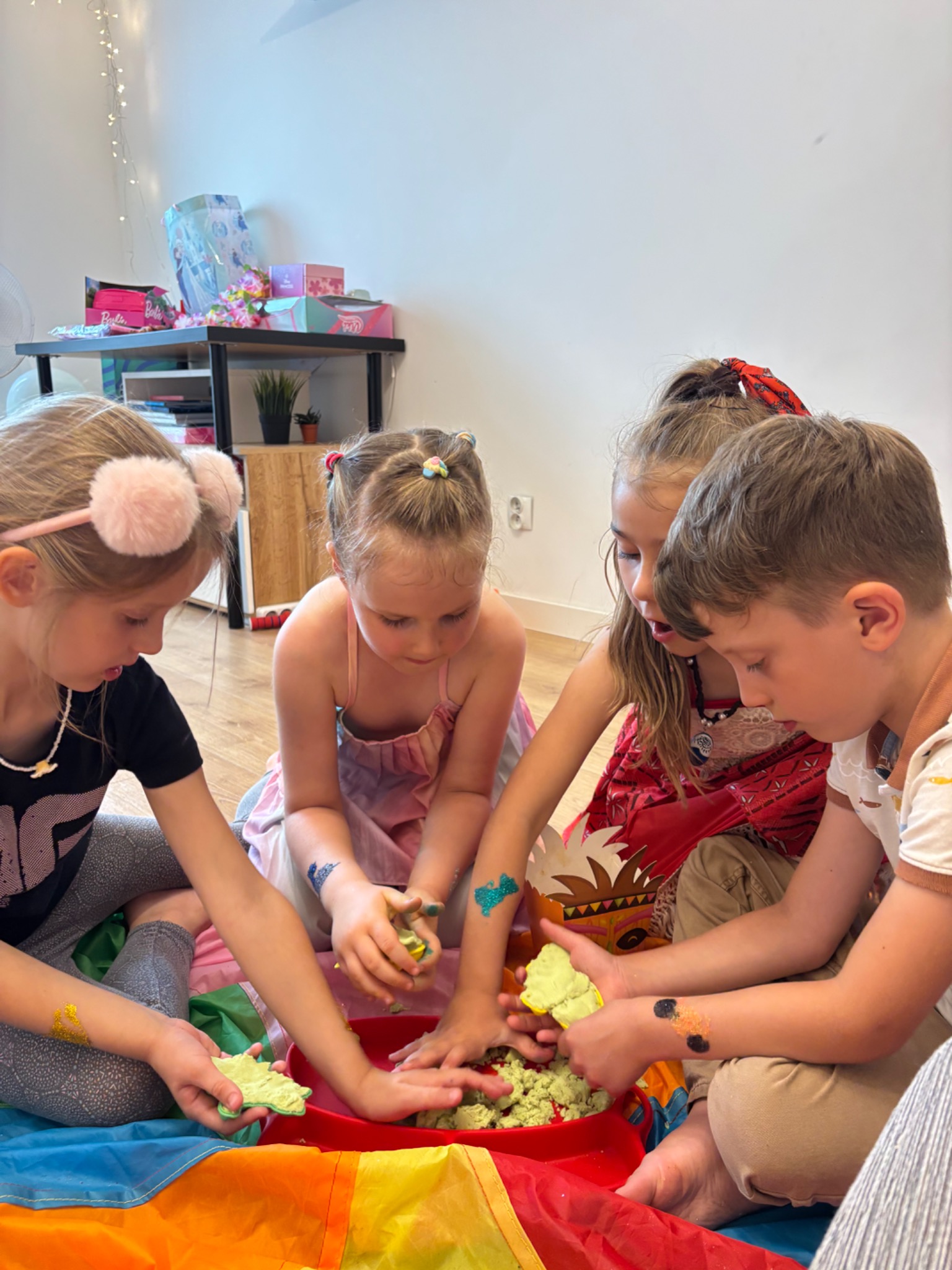 Czterech uśmiechniętych dzieci bawi się kinetycznym piaskiem w czerwonej misce na kolorowej chuście, z brokatowymi tatuażami na ramionach, kreatywna zabawa.