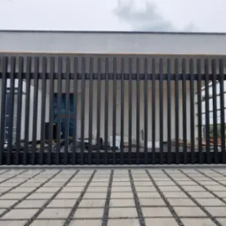 Nowoczesna brama wjazdowa z pionowych, czarnych profili metalowych, osadzona między betonowymi słupkami na tle budynku w trakcie budowy, z widoczną kostką brukową na podjeździe.