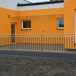 VERME SPÓŁKA Z OGRANICZONĄ ODPOWIEDZIALNOŚCIĄ - Nowoczesna balustrada ze stali nierdzewnej na balkonie budynku o pomarańczowej elewacji i przeszklonym zadaszeniu, widok z zewnątrz na trawnik.