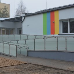 VERME SPÓŁKA Z OGRANICZONĄ ODPOWIEDZIALNOŚCIĄ - Zewnętrzna balustrada ze stali nierdzewnej z matowym szkłem, zabezpieczająca podjazd dla osób z niepełnosprawnościami przy budynku z kolorowymi akcentami.