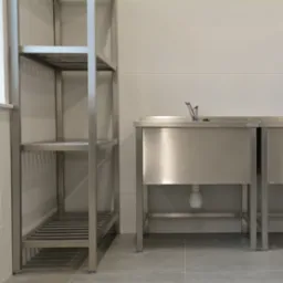 Wnętrze z metalową umywalką z kranem i półką na tle białych płytek. Minimalistyczny design, chłodna kolorystyka, widoczny syfon. Ujęcie z dołu.