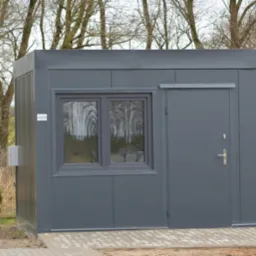 Szara, metalowa budka z oknem i drzwiami, stojąca na kostce brukowej. W oknie odbijają się drzewa. Widoczne panele ścienne i skrzynka elektryczna na ścianie.