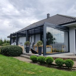 VERME SPÓŁKA Z OGRANICZONĄ ODPOWIEDZIALNOŚCIĄ - Szklana zabudowa tarasu z czarną ramą i widocznym wnętrzem z meblami ogrodowymi. Na tarasie pies, w tle dom z szarą dachówką i zielony trawnik.