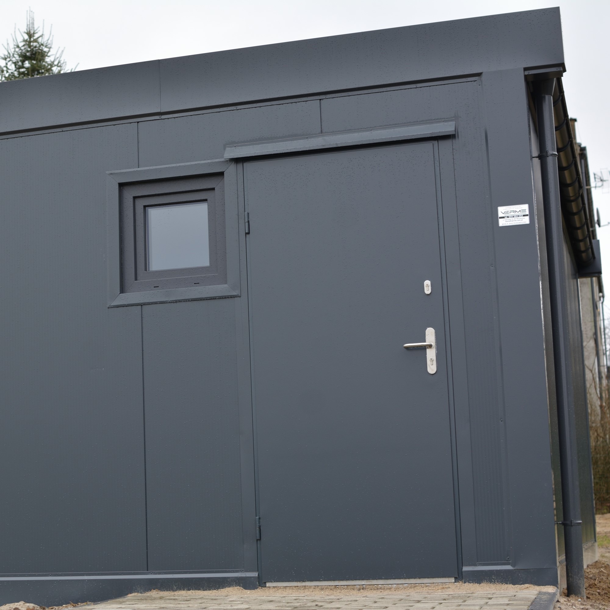 Nowoczesny, grafitowy pawilon z oknem i drzwiami, wykończony na zewnątrz metalowymi panelami. Widoczne detale wykończeniowe i fragment kostki brukowej u podstawy.