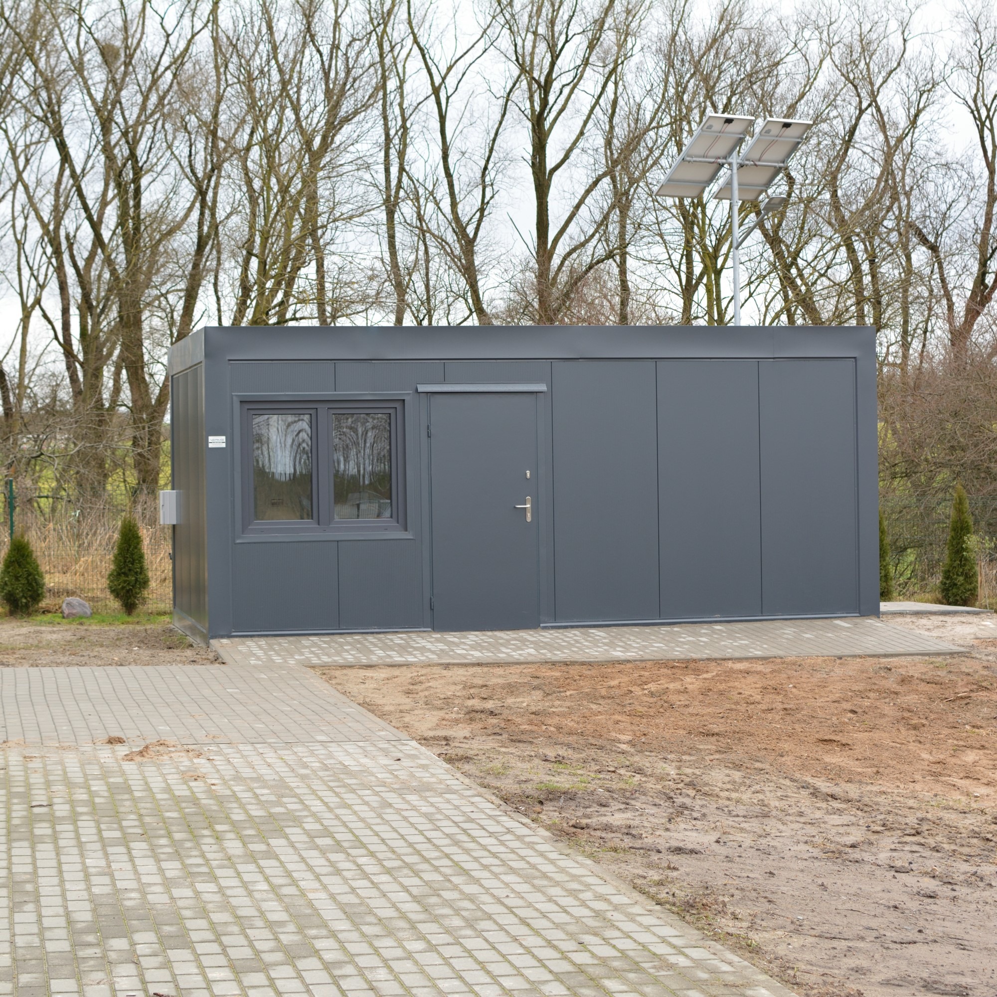 Szary pawilon modułowy z oknem i drzwiami, oświetlony panelami solarnymi, na tle drzew bez liści i chodnika z kostki brukowej. Widok z zewnątrz, dzień.