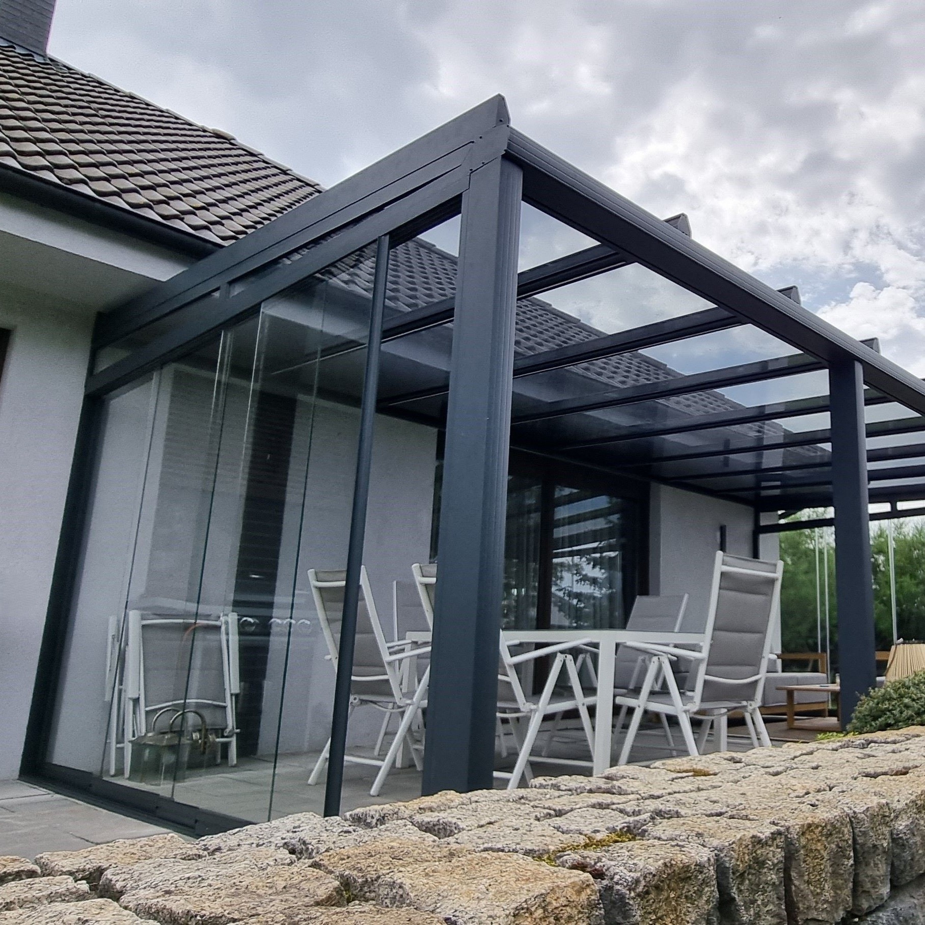 Nowoczesna pergola z metalową konstrukcją i szklanym dachem, przylegająca do domu. Widoczne meble ogrodowe i kamienne wykończenie. Szare niebo w tle.