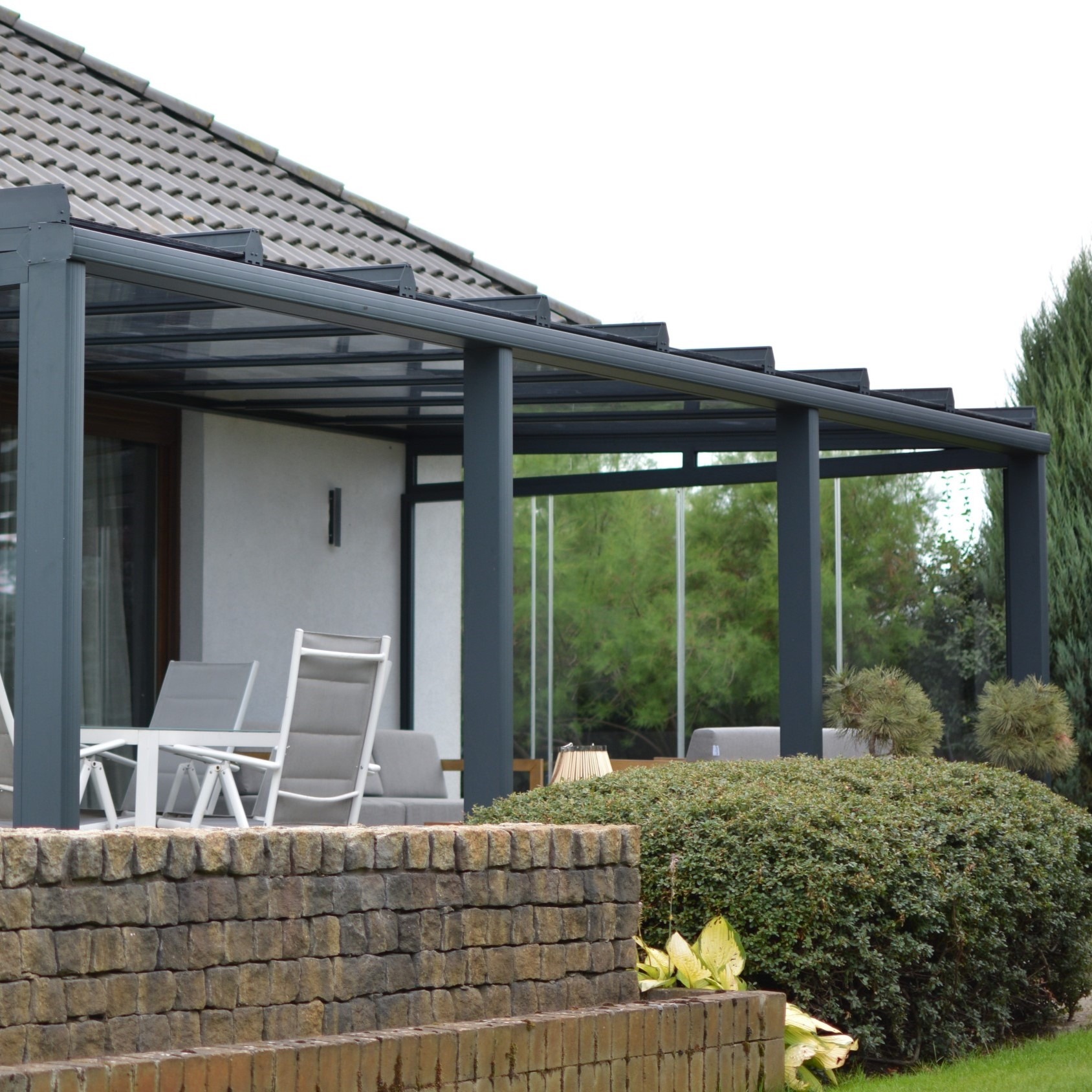 Nowoczesna pergola metalowa z przeszklonym dachem, szare słupy, widok z zewnątrz na taras z meblami ogrodowymi, kamienny murek na pierwszym planie.