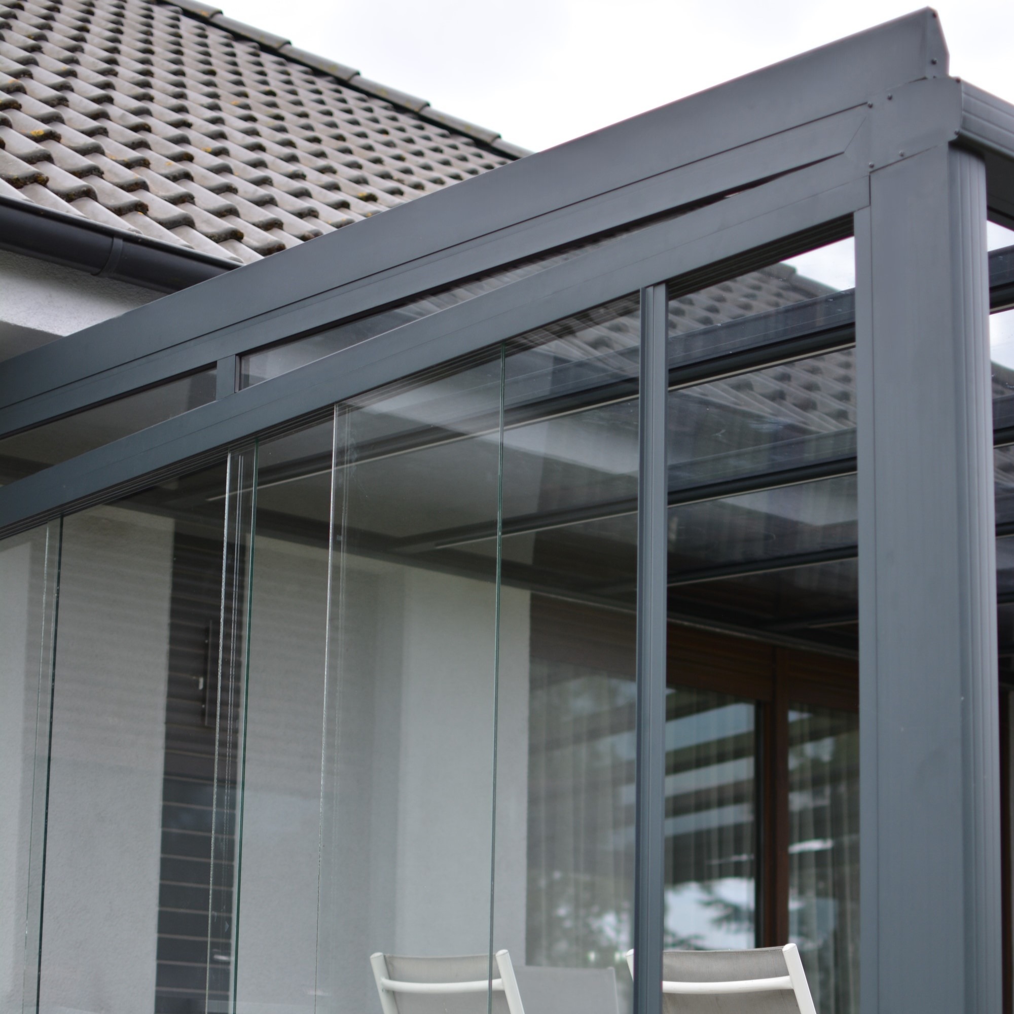 Szklana zabudowa balkonu z szarą, metalową konstrukcją, widoczna z zewnątrz. W tle fragment dachu domu z szarą dachówką. Częściowo widoczne krzesła.
